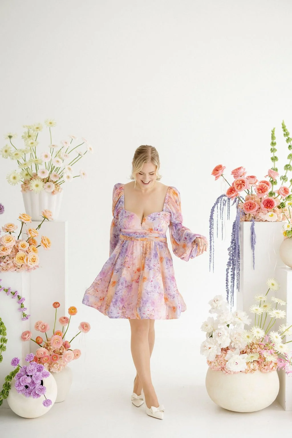 A woman smiling and dancing in a floral dress surrounded by colorful flower arrangements in white vases against a white background.