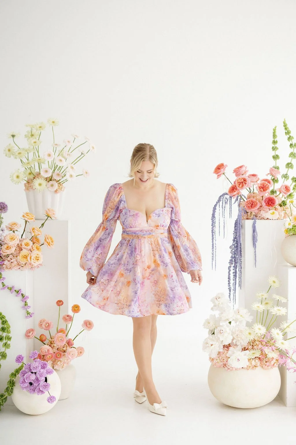 A woman in a pastel floral dress standing amidst large vases filled with pink, purple, and white flowers in a bright, white studio.