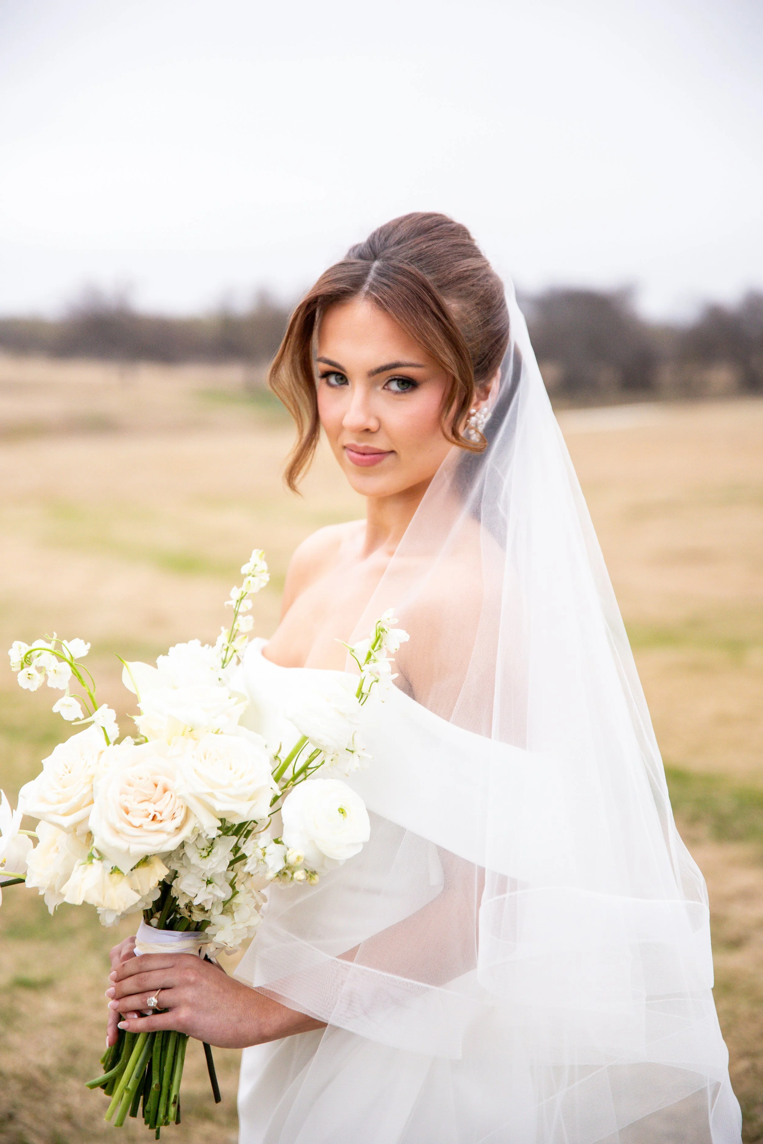 A bride with styled brown hair, wearing a white wedding dress and veil, holding a bouquet of white and light pink flowers, standing outdoors with blurred trees and grass in the background.