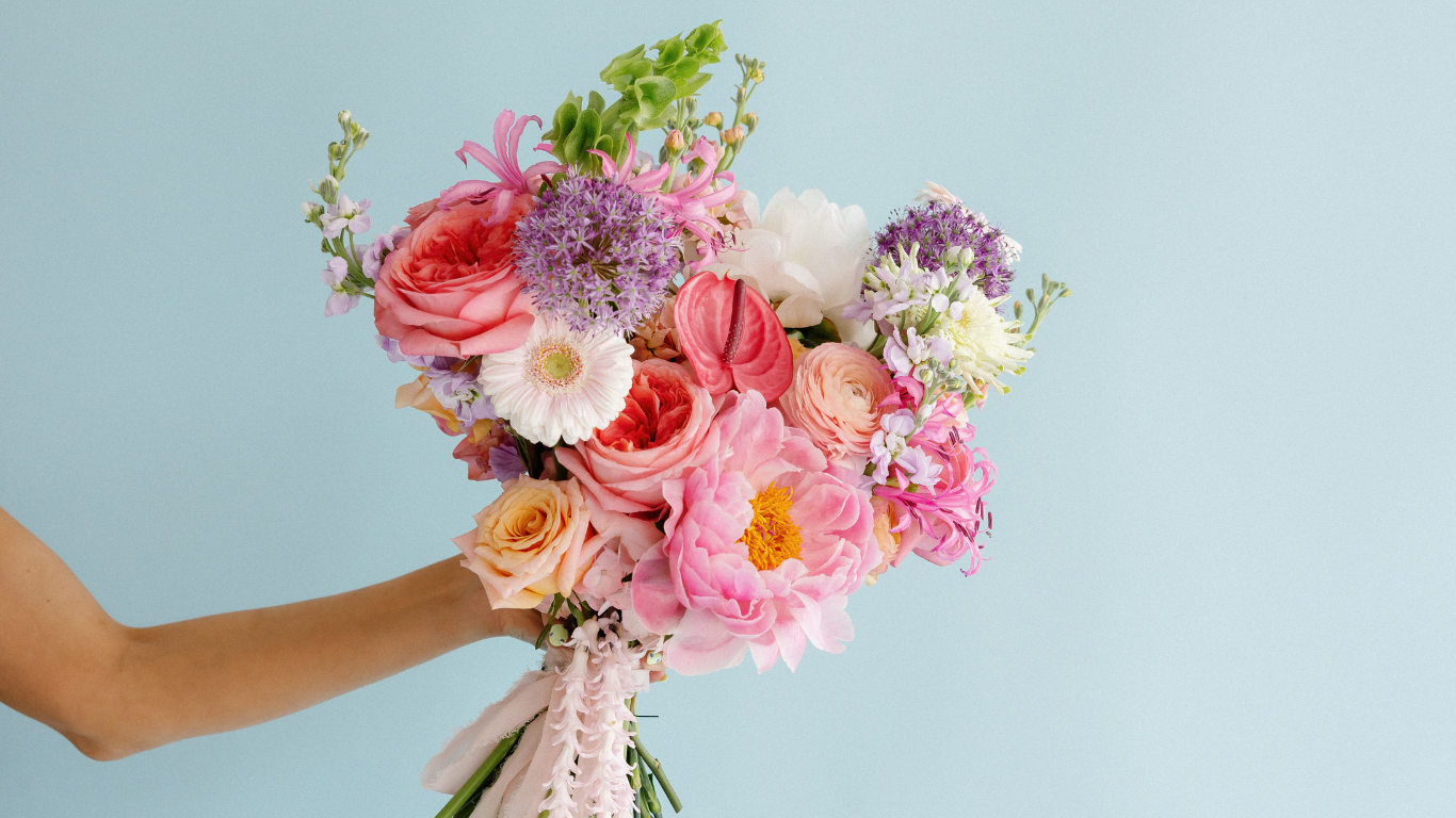 A person holding a bouquet of colorful flowers including roses, peonies, daisies, and other blooms against a light blue background.