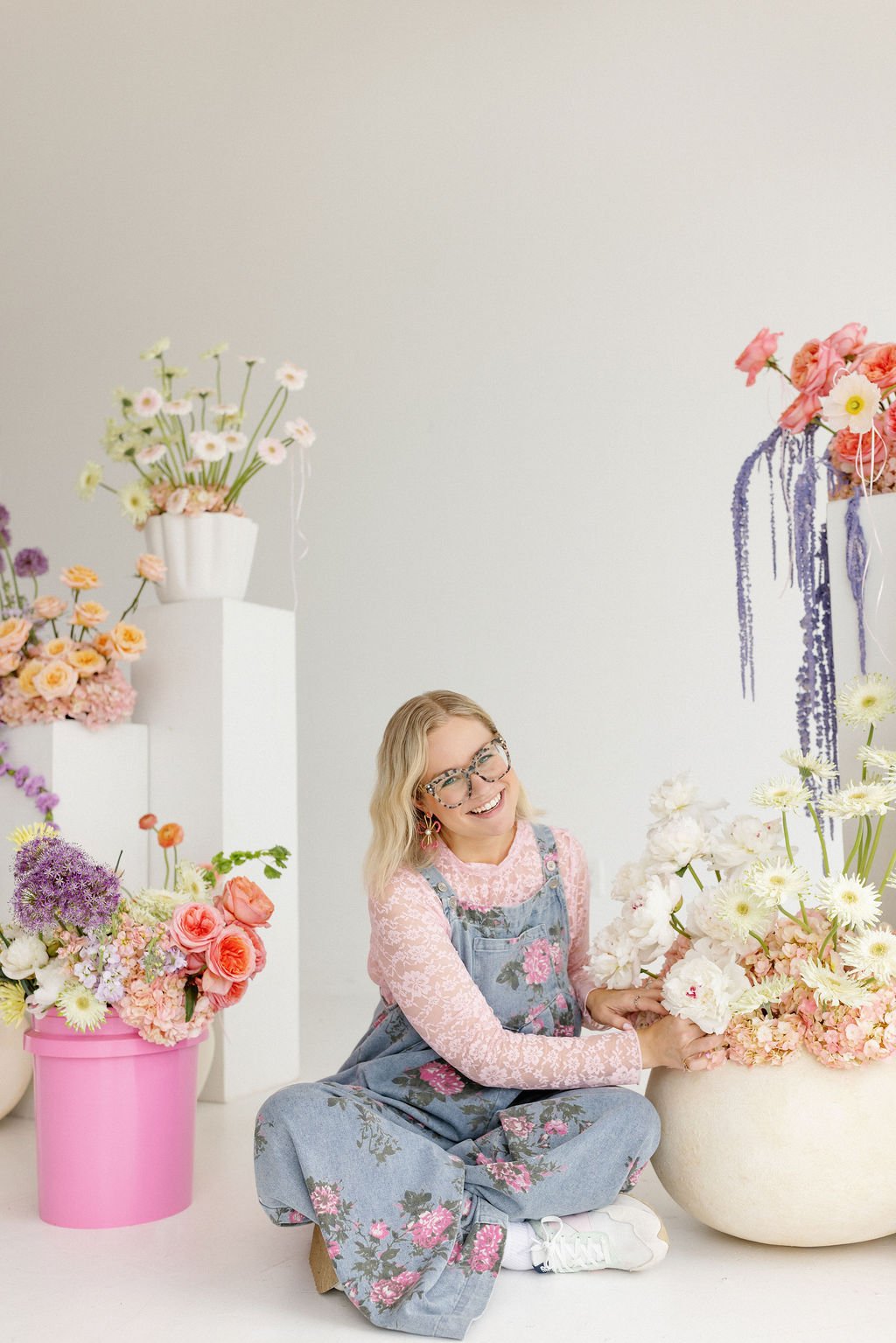 A smiling woman with glasses and blonde hair, wearing a pink lace top and floral overalls, sitting on the floor and arranging white and pink flowers in a large beige vase, surrounded by colorful flower arrangements in various vases and pots.