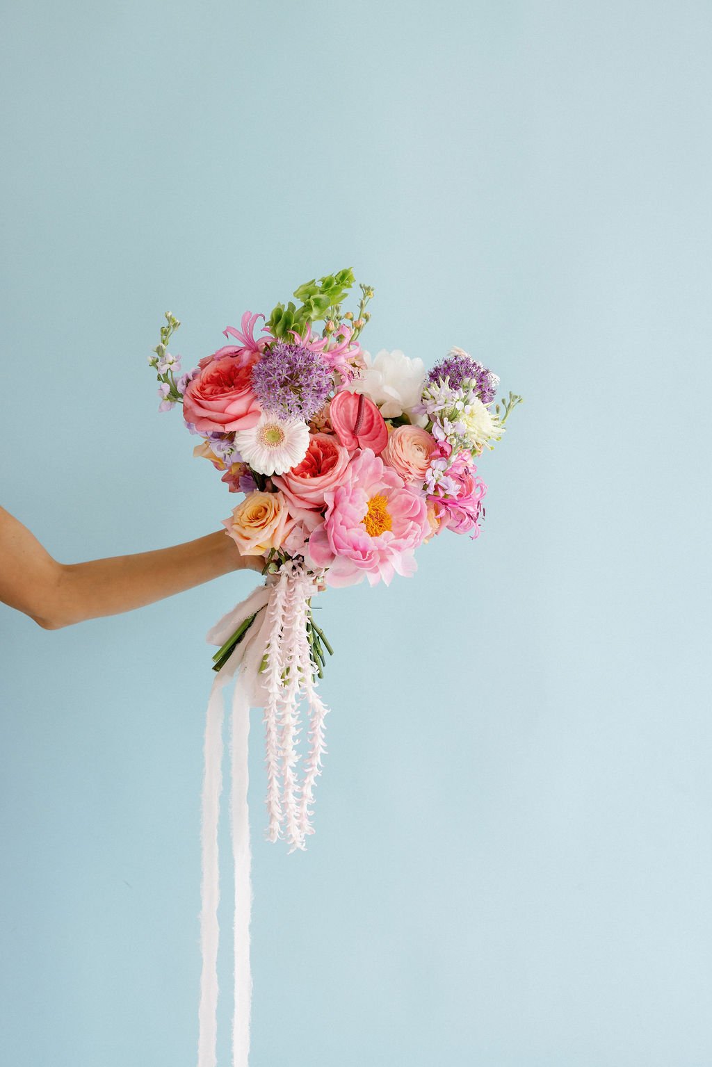 A person holding a bouquet of pink, purple, white, and peach flowers against a light blue background.