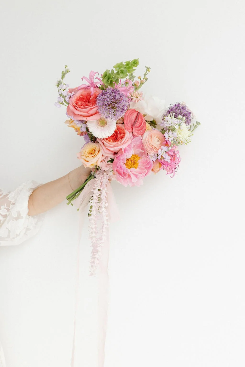 A hand holding a colorful bouquet of various pink, purple, peach, and white flowers against a plain white background.