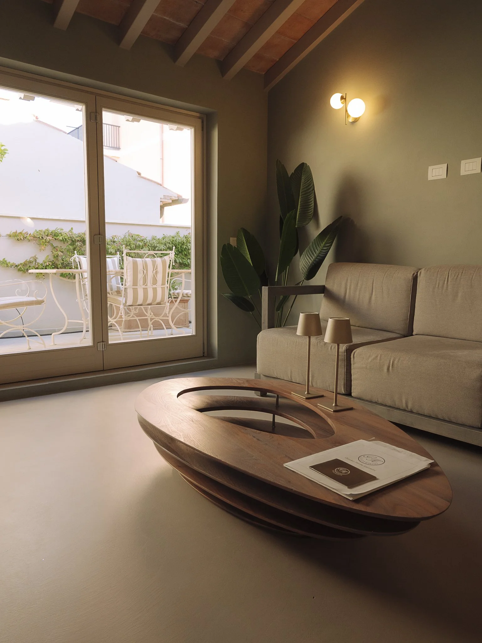 Living room with beige sofa, two small beige lamps on a wooden coffee table, large green plant next to the window, and outdoor balcony with white wrought iron furniture and greenery.