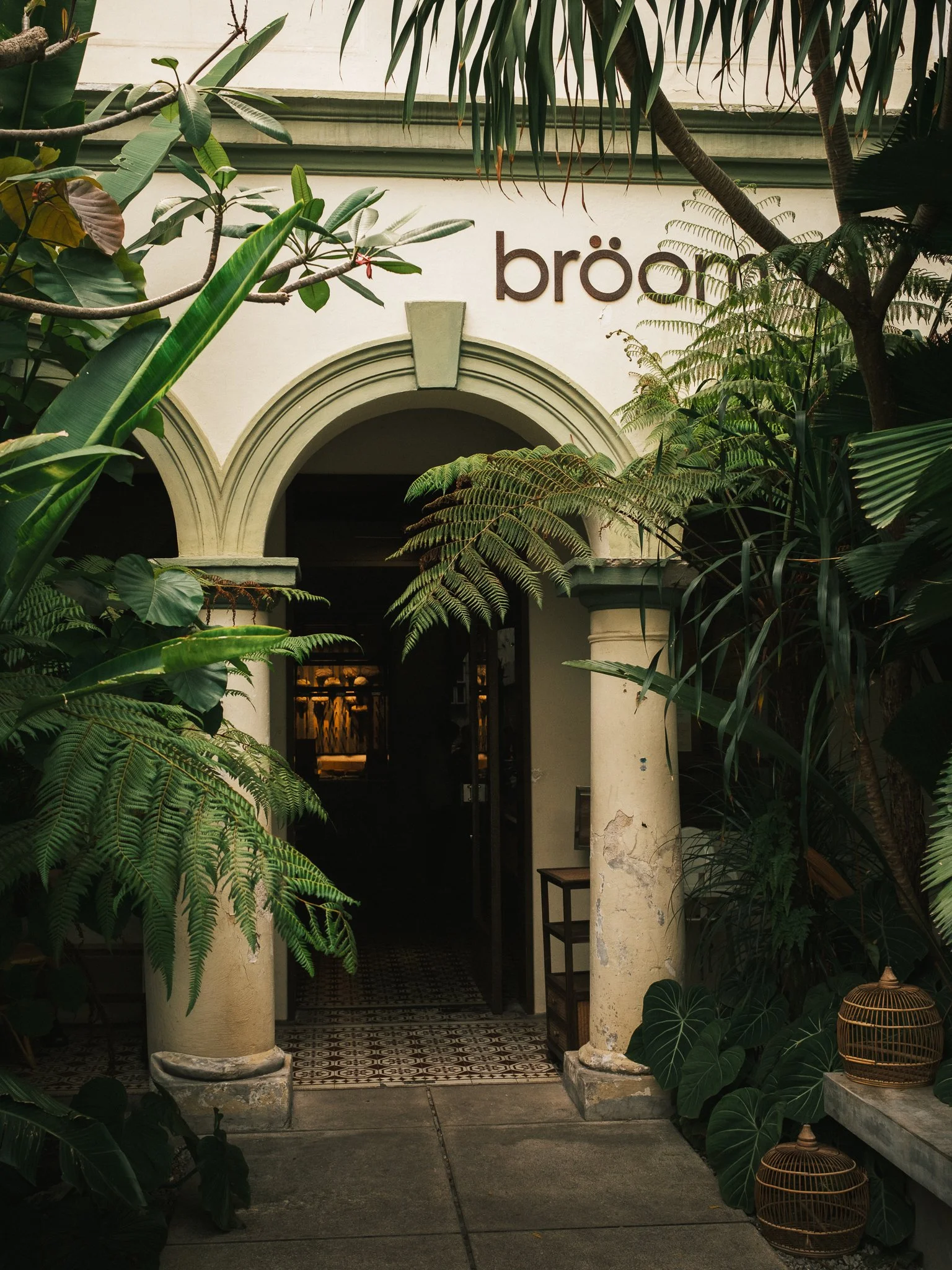 Entrance way with two large cream-colored columns, surrounded by lush green plants, with the word 'brea' on the wall above. There are plants hanging overhead and placed beside the columns, with two rattan baskets on the right.