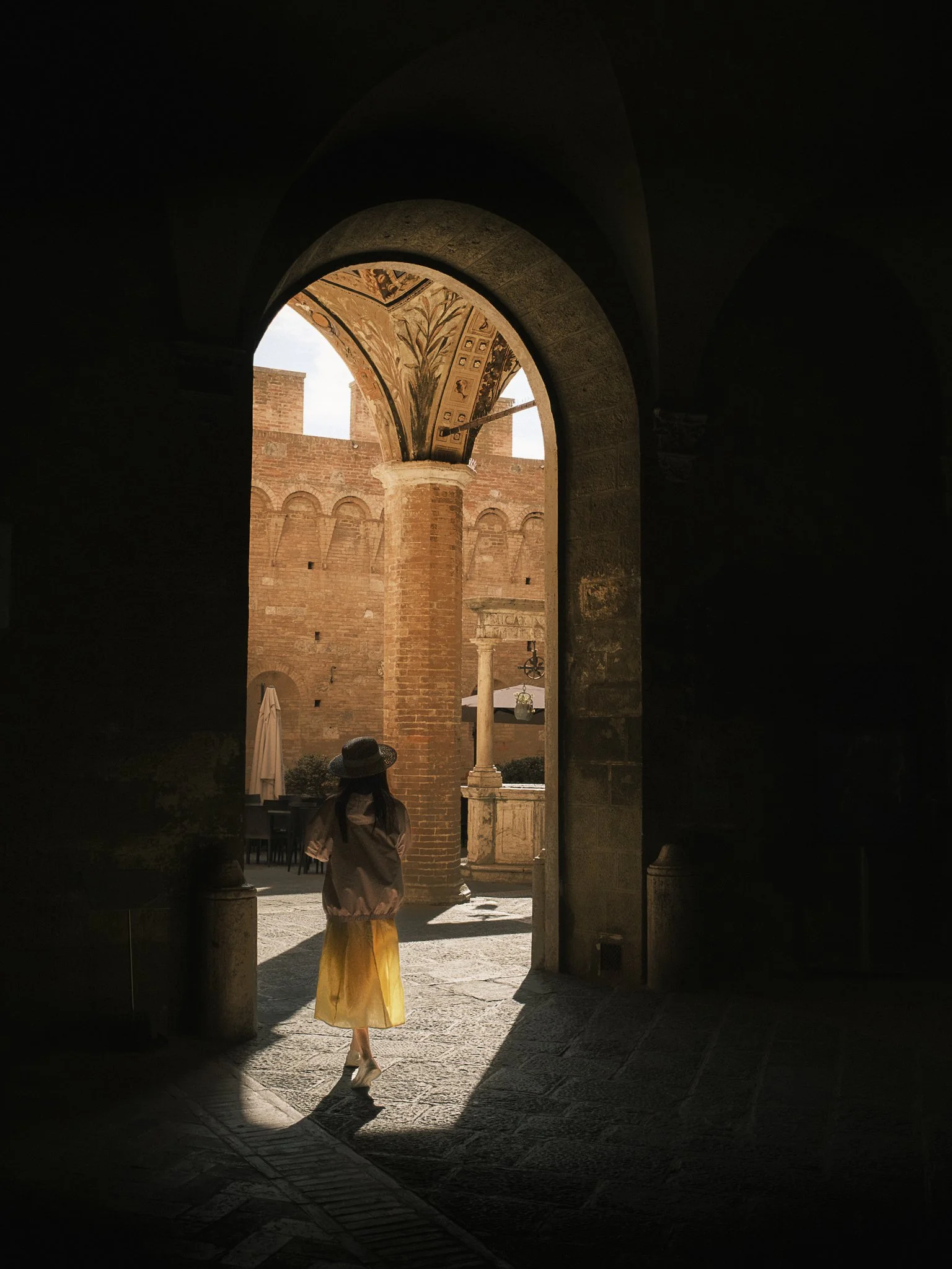 A person in a yellow skirt and white top walking through a dark arched doorway into a sunlit courtyard with brick walls and classical architecture.