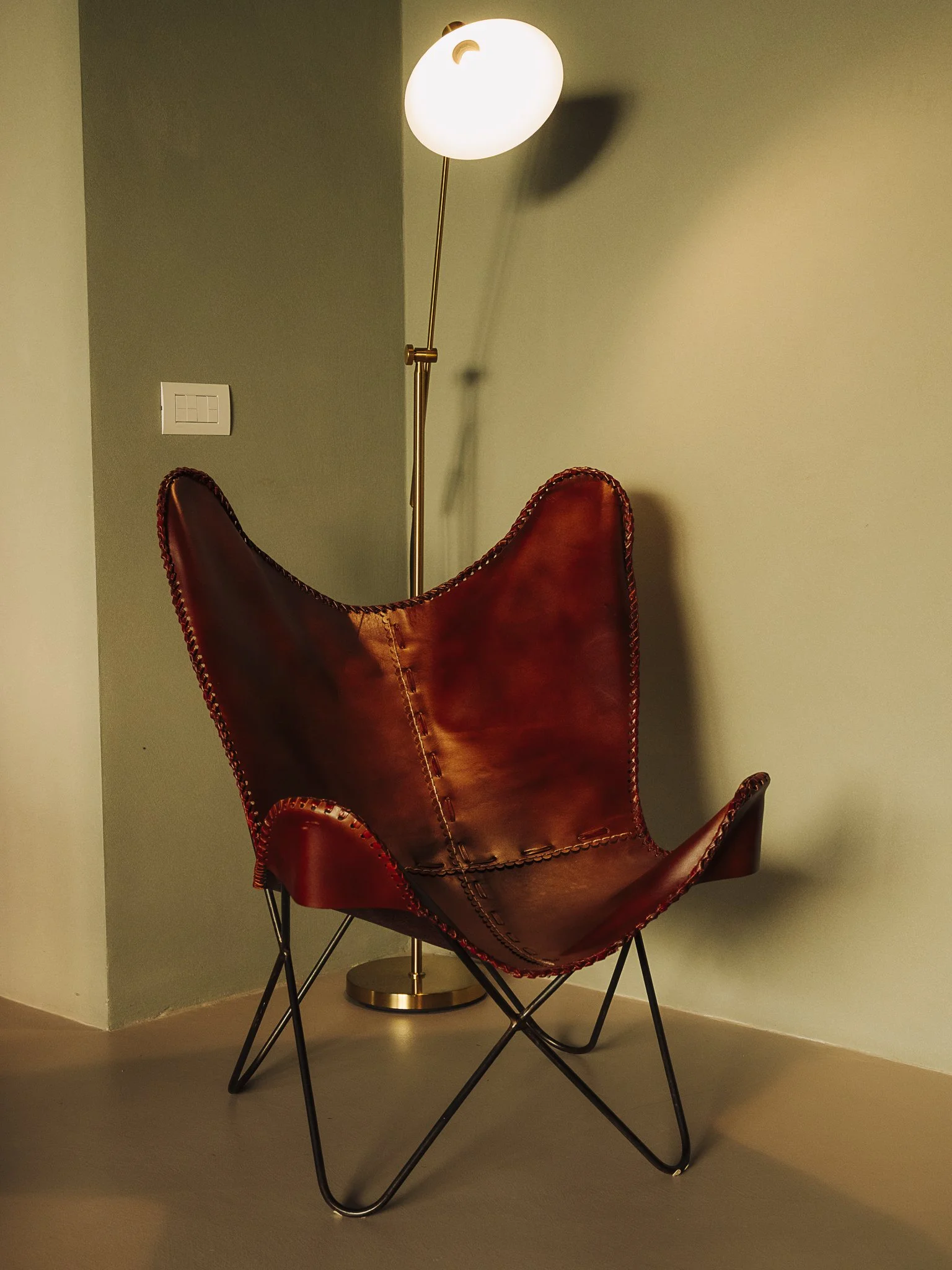 A red leather butterfly-shaped butterfly chair with black metal legs, positioned in front of a green wall and a white wall corner, illuminated by a white globe floor lamp.