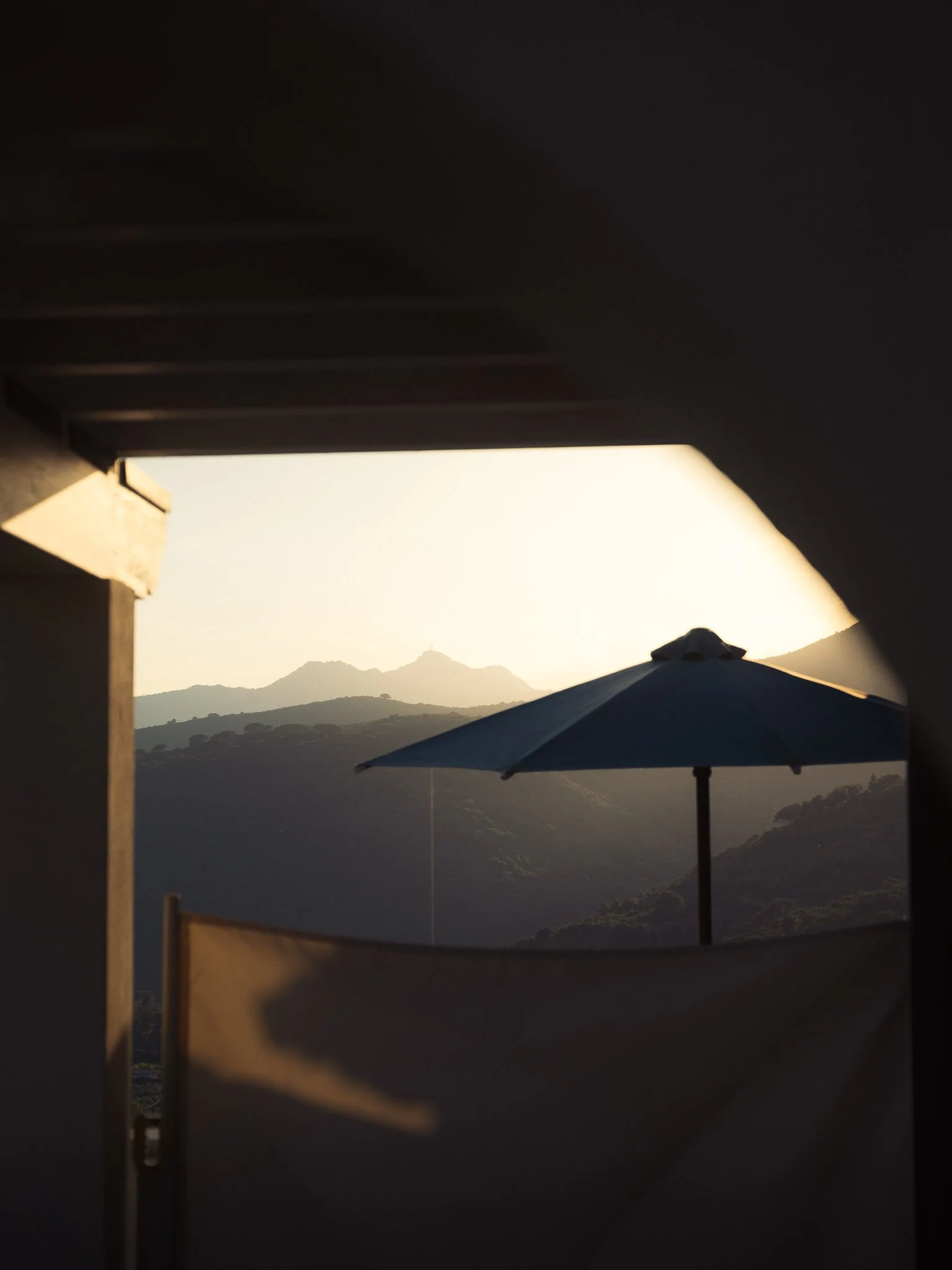 Sunset view through a window or opening showing mountains, with a blue umbrella and patio umbrella in the foreground.