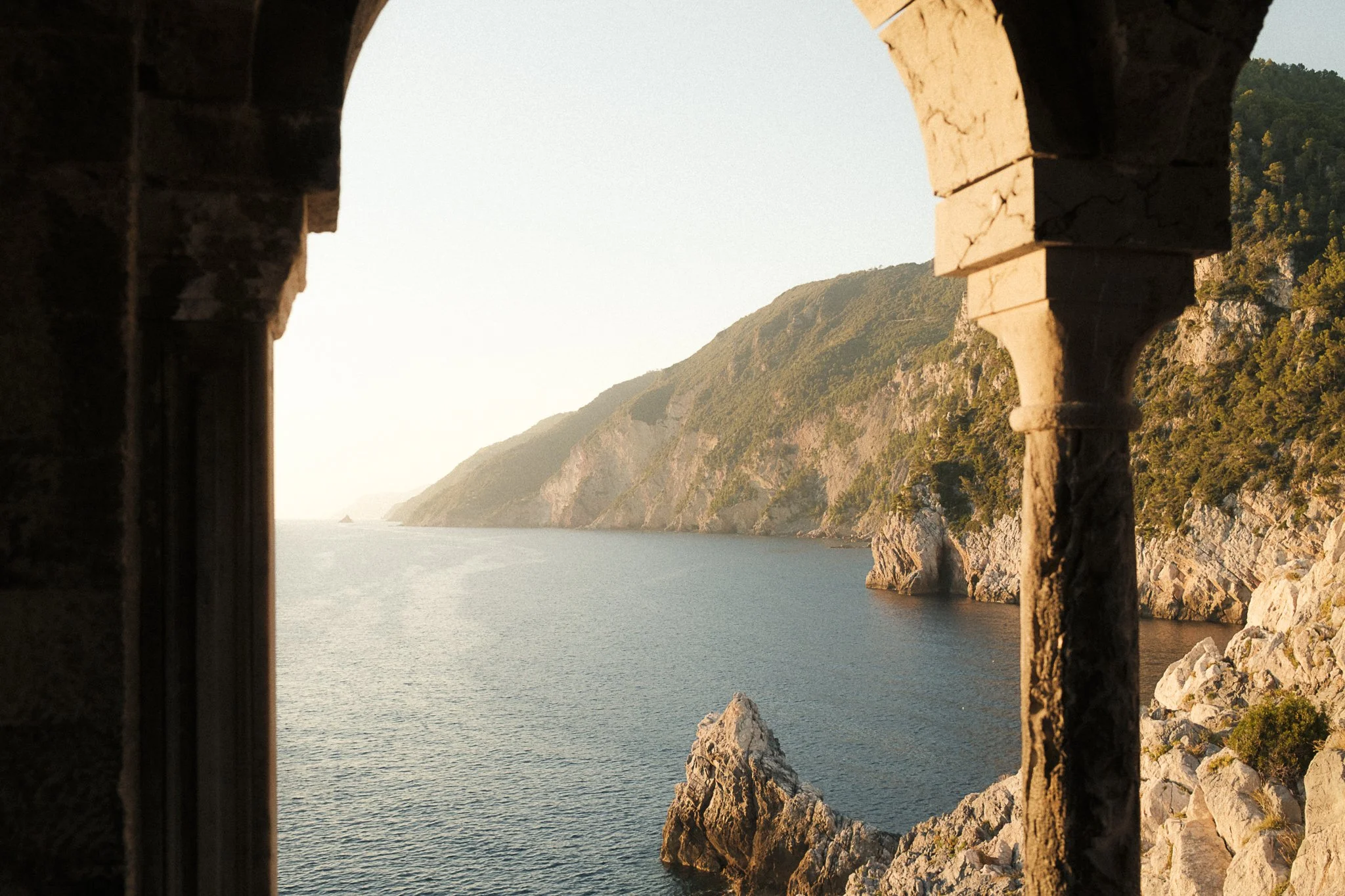 Scenic coastal view through an arched stone window, with green cliffs and a calm body of water in the distance.