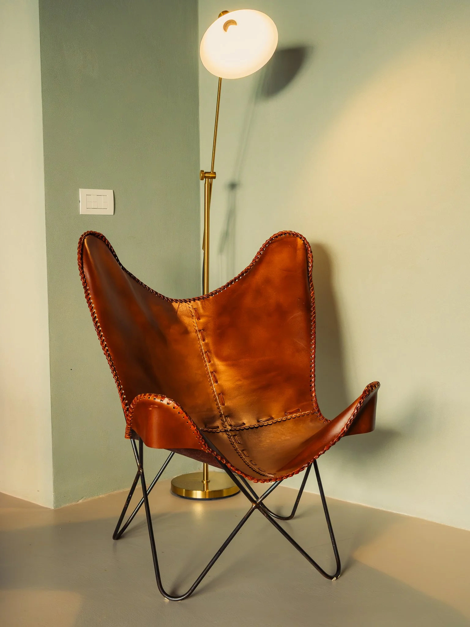 A modern leather butterfly chair with a black metal frame and a tall, curved brass floor lamp next to it, against light green and off-white walls.