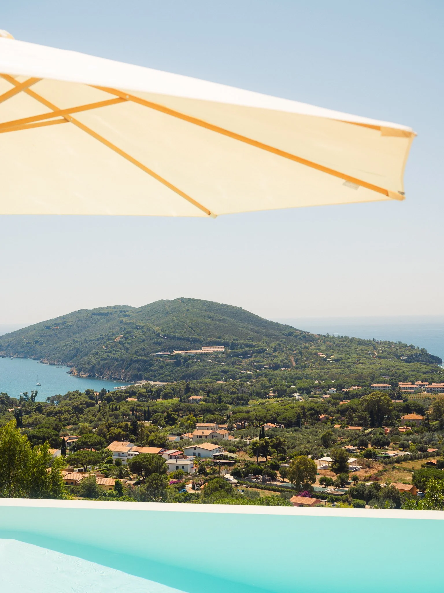 Scenic view of a coastal hillside town with many houses, lush green trees, and a body of water in the distance, partially covered by a yellow and white beach umbrella.