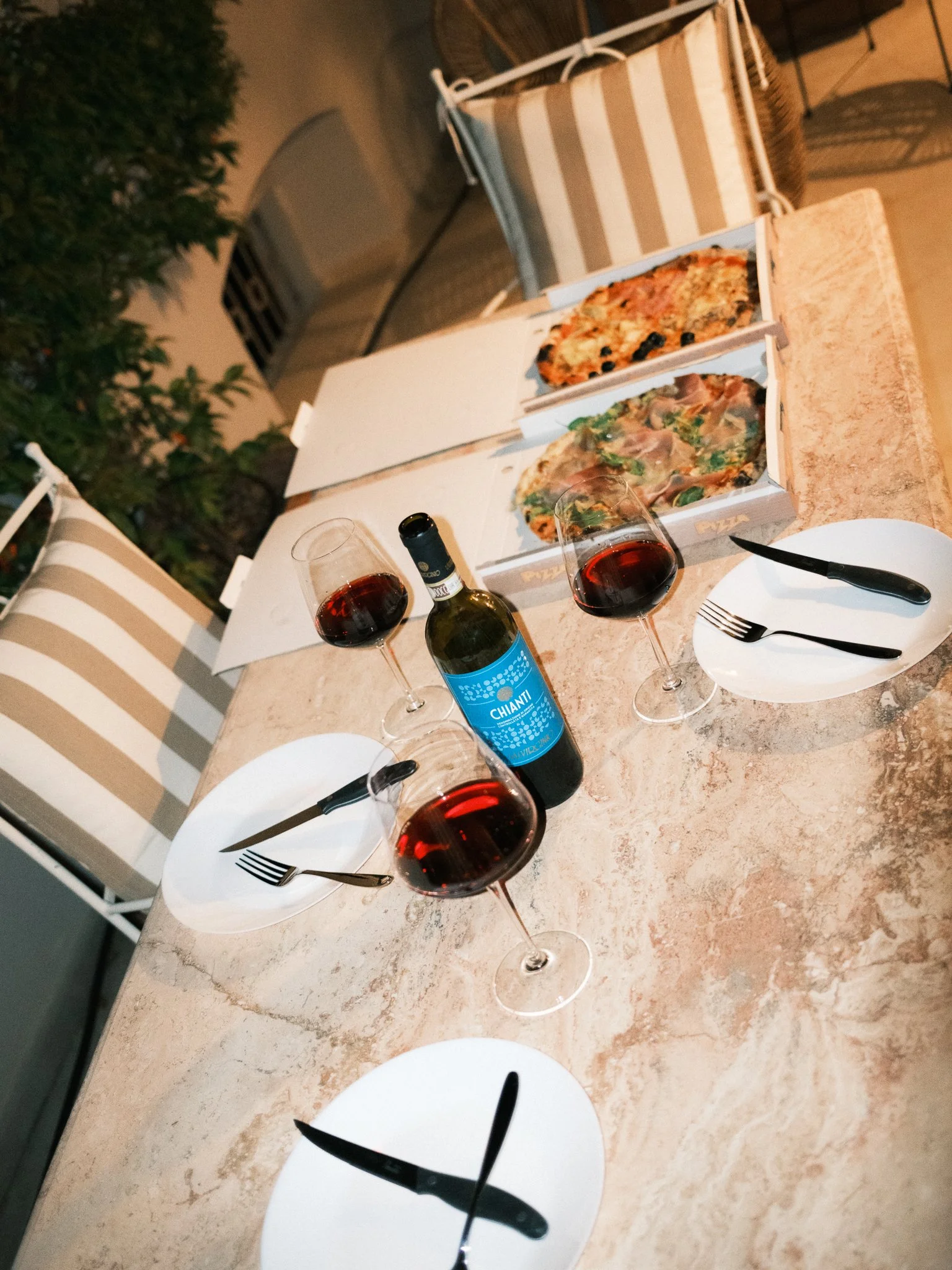 Two glasses of red wine on a table with pizza boxes, plates, and cutlery, likely set for a meal, against a background of outdoor patio furniture and plants at night.