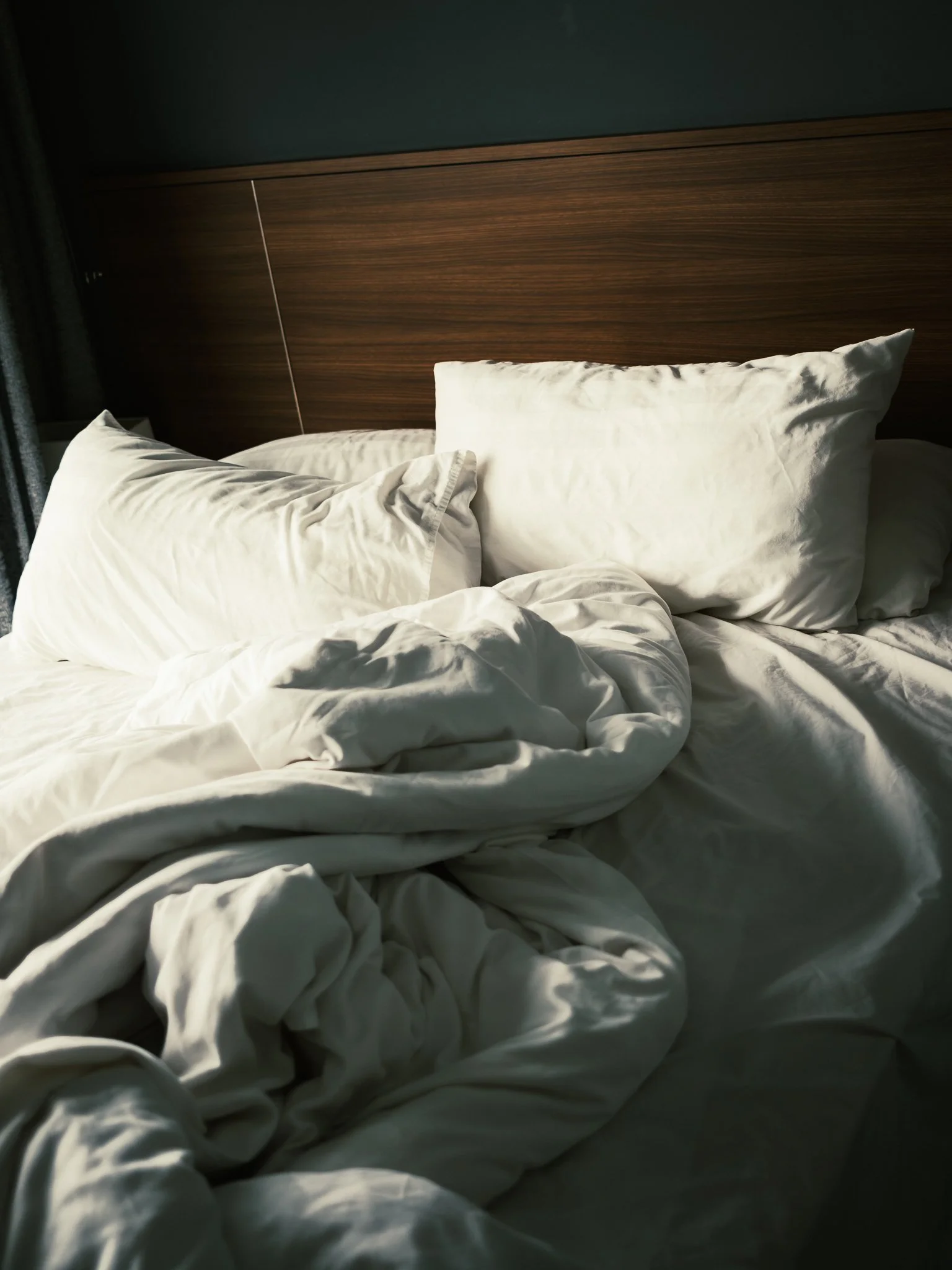 A bed with rumpled white sheets and pillows, a wooden headboard, part of a dark wall, and a window with curtains on the left side.