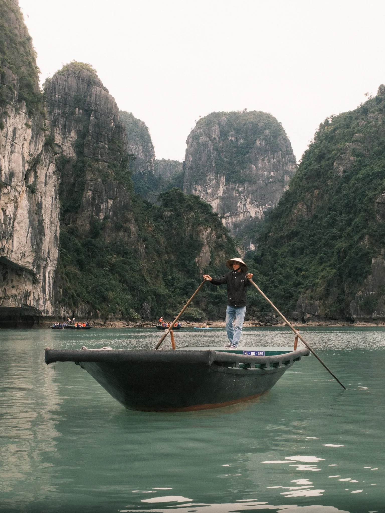A person standing on a traditional boat in Ha Long Bay surrounded by tall, lush green mountains with rocky cliffs in the background, wearing a conical hat and black jacket.