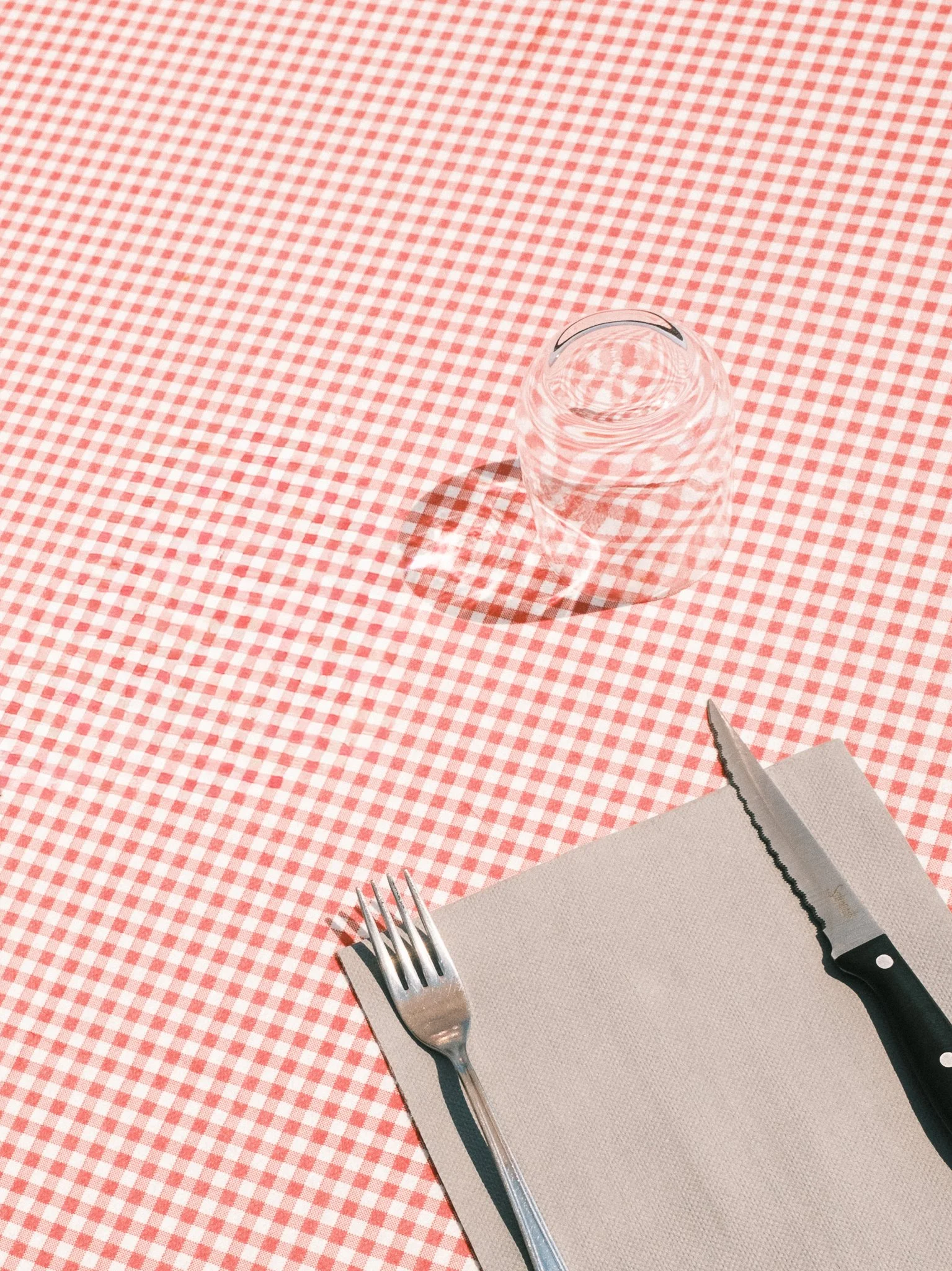 A transparent glass of water on a red and white checkered tablecloth, with a fork, knife, and napkin nearby.