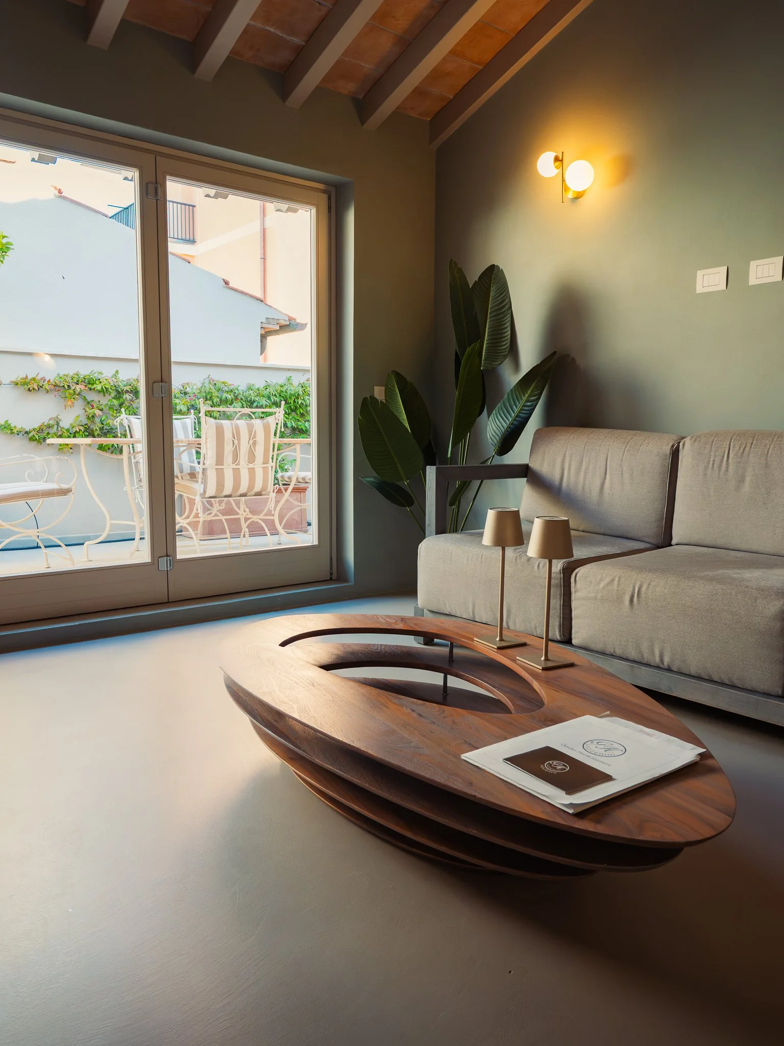 A living room with a sliding glass door leading to a balcony with outdoor chairs and green plants. Inside, there is a beige sofa, a wooden coffee table with a magazine, and a tall green plant. A wall sconce light is on, illuminating the wall with swi