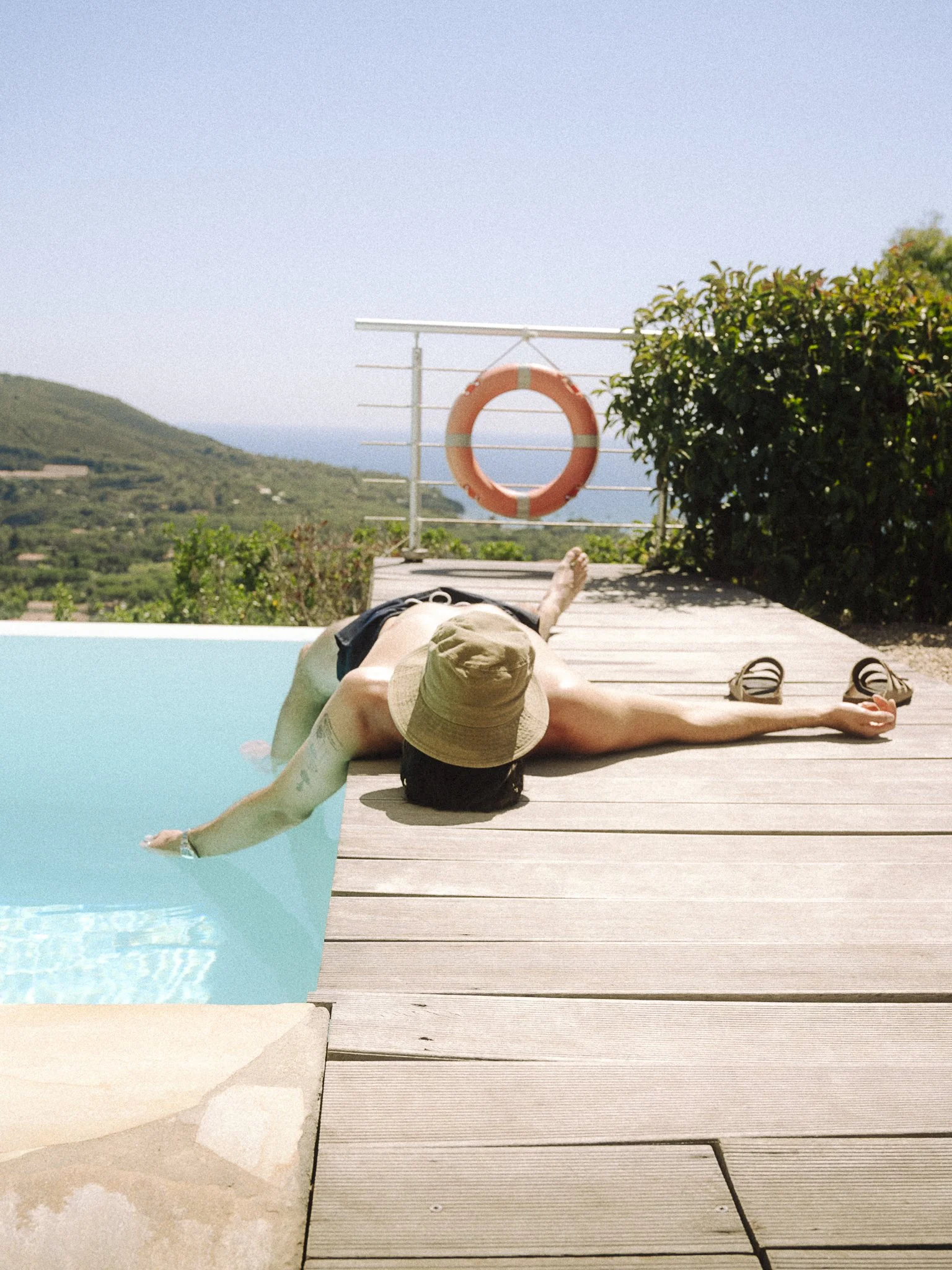 Person lying on a poolside wooden deck, wearing a sun hat, with an arm stretched out toward the water, beside a pair of sandals, overlooking a scenic hilly landscape and the ocean in the background.