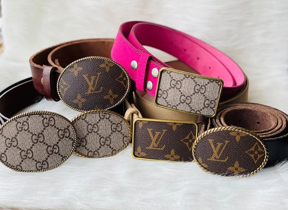 Collection of designer belts and belt buckles with Louis Vuitton logos, featuring pink, brown, beige, and green colors, arranged on a white surface.