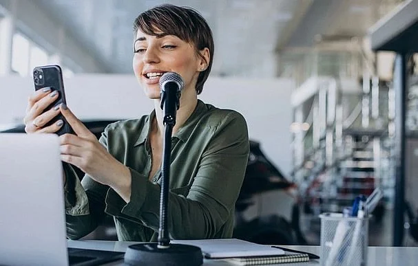 Woman speaking into microphone while looking at her phone in a modern office or studio setting.