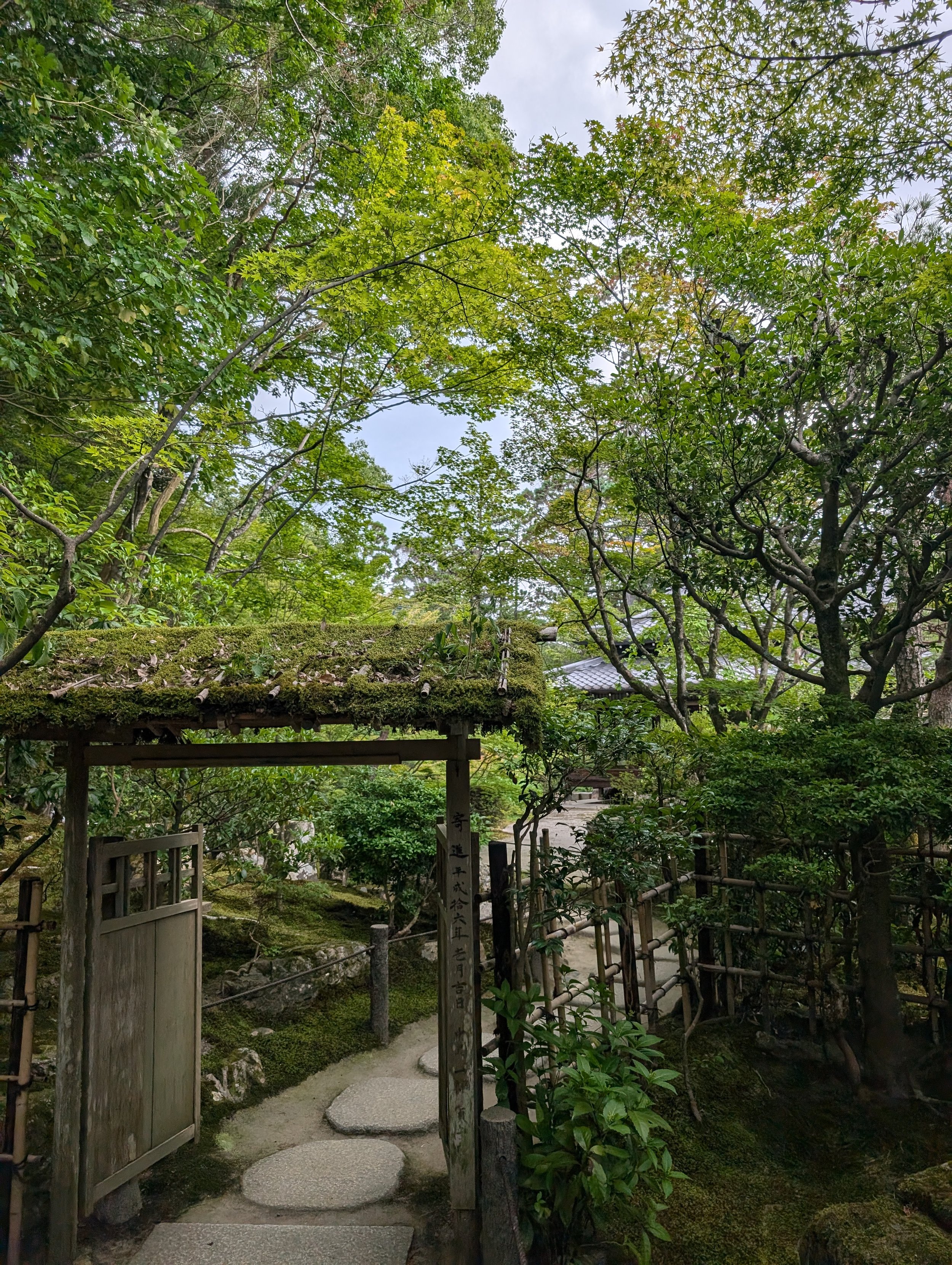 A traditional Japanese garden scene with a moss-covered wooden gate, stone pathway, and lush green trees and foliage.