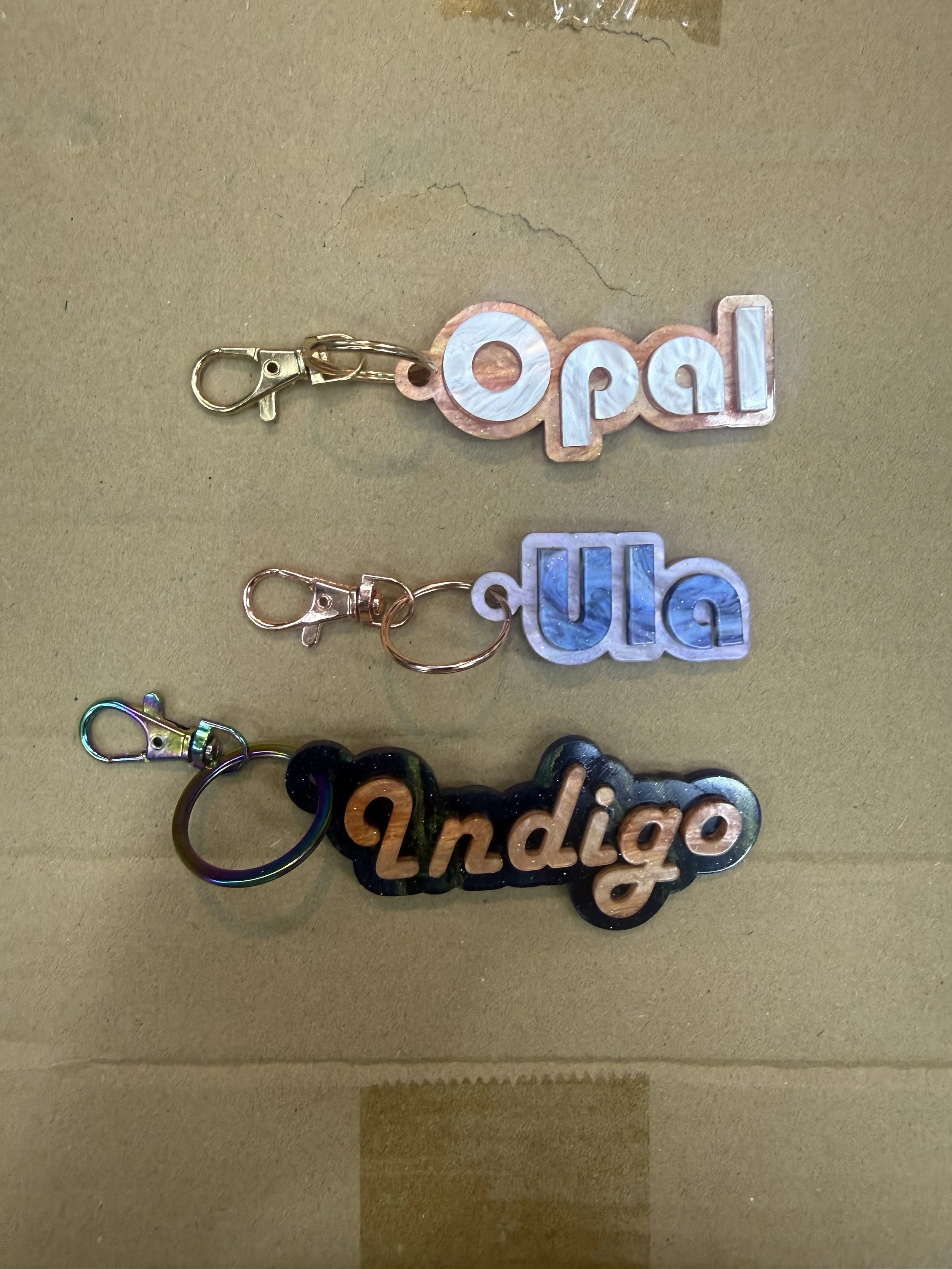 Colorful keychains with the words 'apol' and 'indigo' written in decorative fonts, attached to metal key rings.