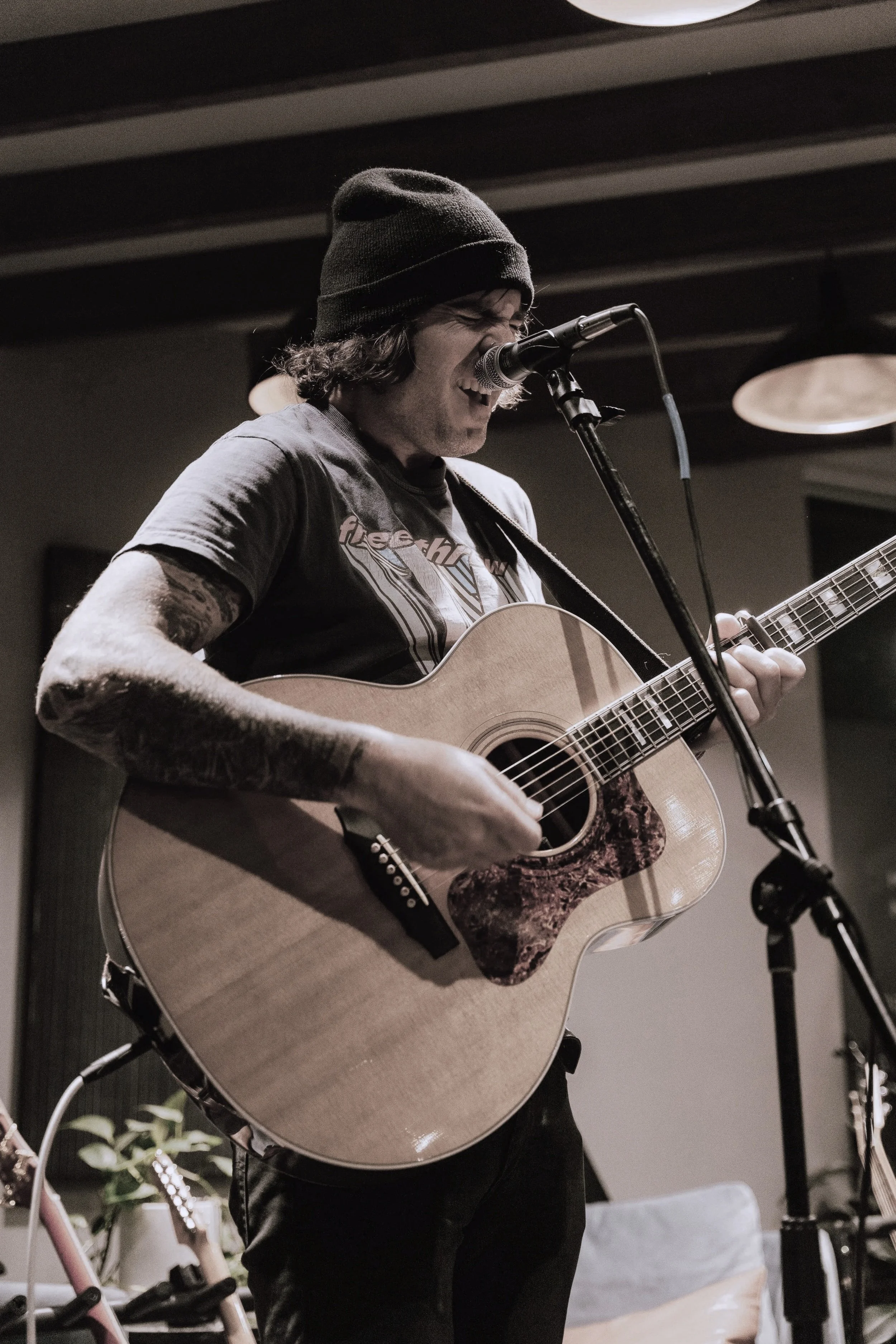 A person wearing a dark beanie hat and a gray T-shirt is playing an acoustic guitar and singing into a microphone indoors.