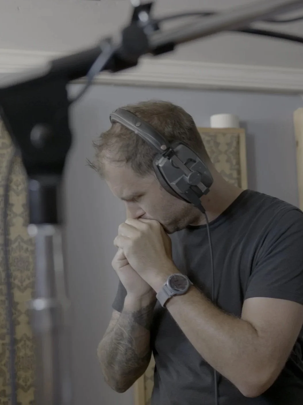 A man with headphones and a watch on his left wrist is praying with his hands clasped in front of his face, eyes closed, in a room with a decorative curtain and a gray wall.