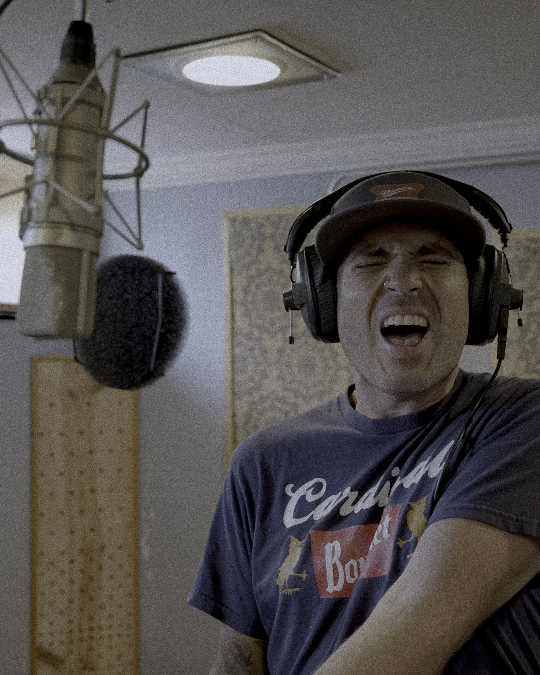 A man wearing a trucker hat and headphones is singing or shouting into a microphone in a recording studio.