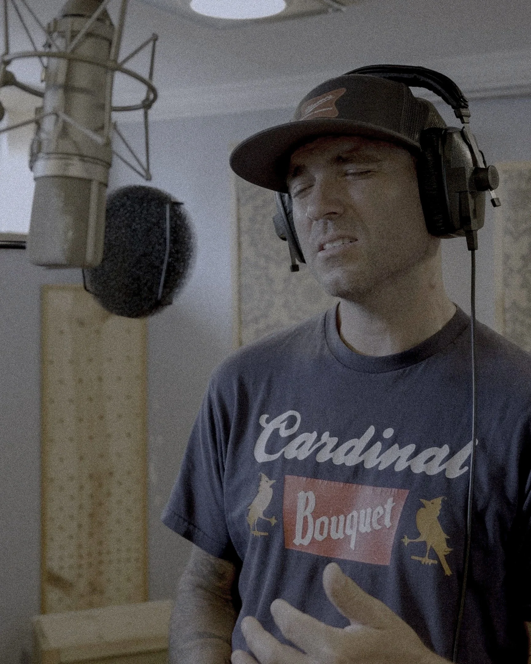 A person wearing a cap and headphones in a recording studio, with a microphone and pop filter in front of him, and wearing a T-shirt that says 'Candy Bouquet'.
