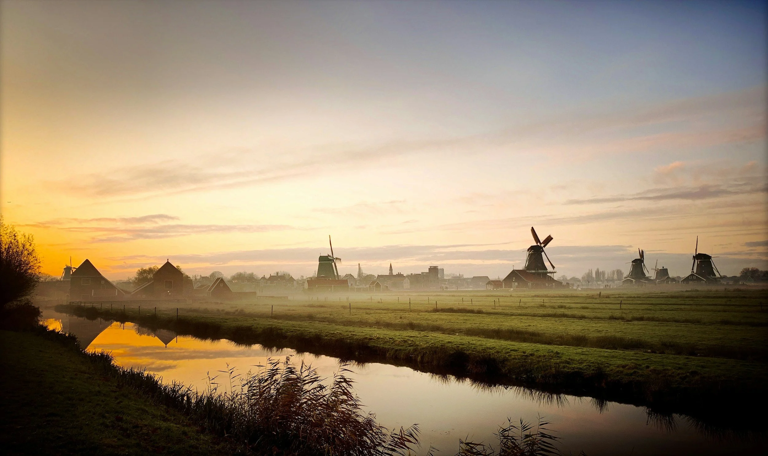 Sunrise over a Dutch landscape with windmills, water canal, green fields, and a few buildings in the distance.