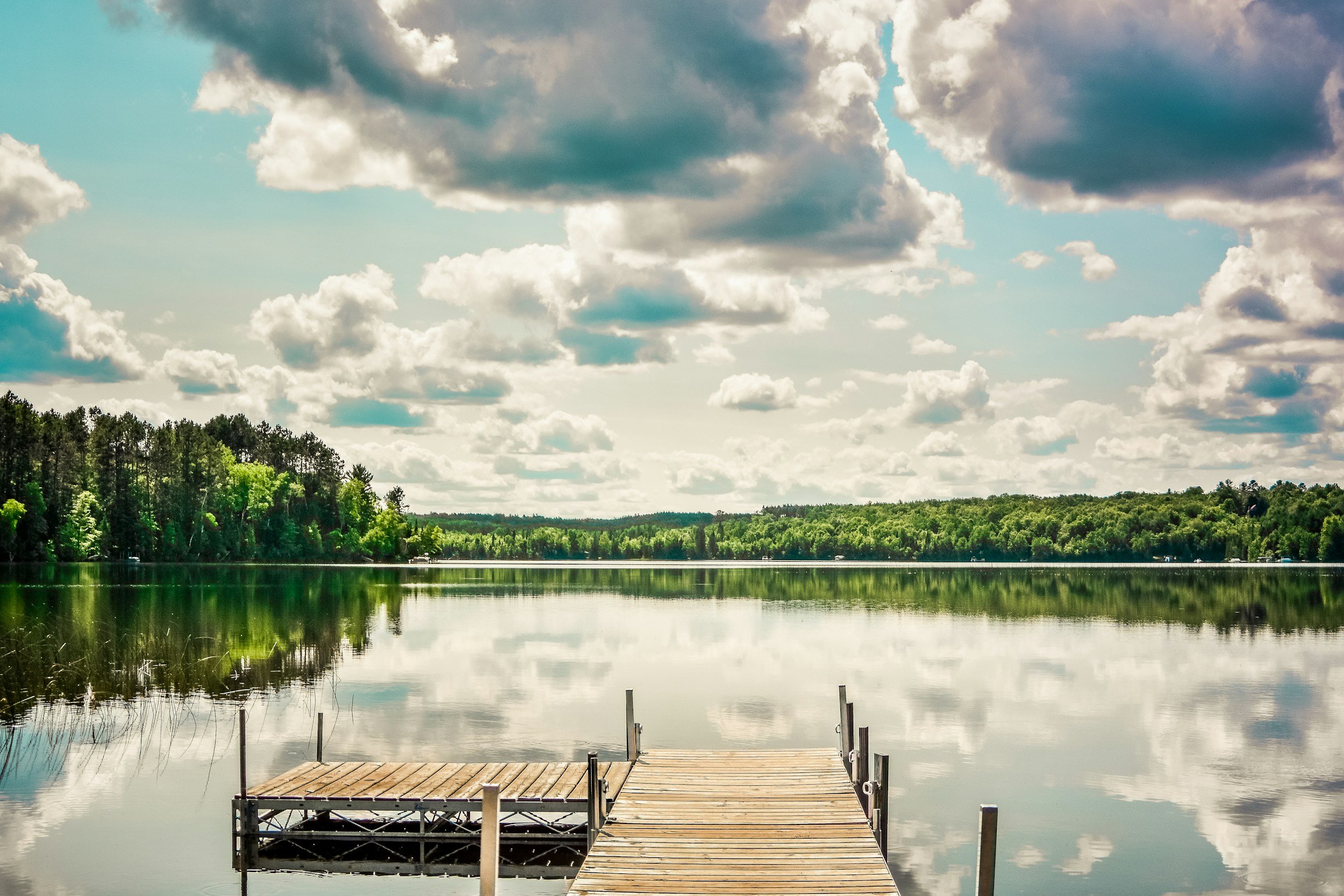 A wooden dock extending into a calm lake, surrounded by green trees and a partly cloudy sky reflected in the water.
