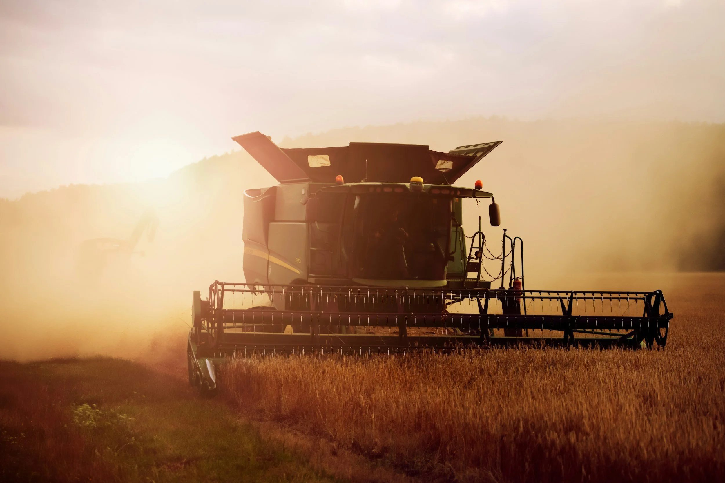 A combine harvester working in a field at dusk or dawn with a bright sun, casting a warm glow and creating a silhouette effect