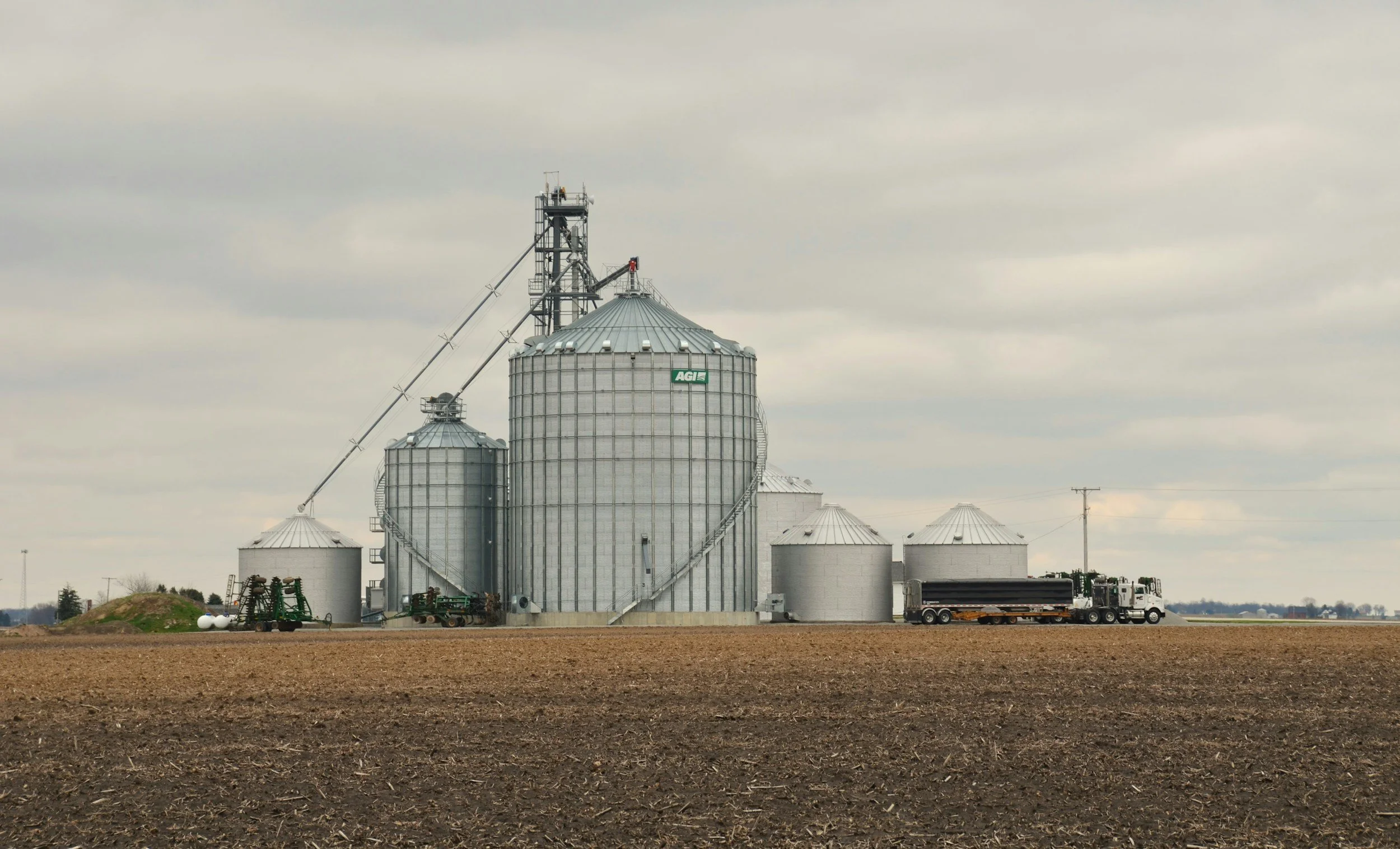 Multiple grain silos on a farm with a truck in front, set against a cloudy sky.