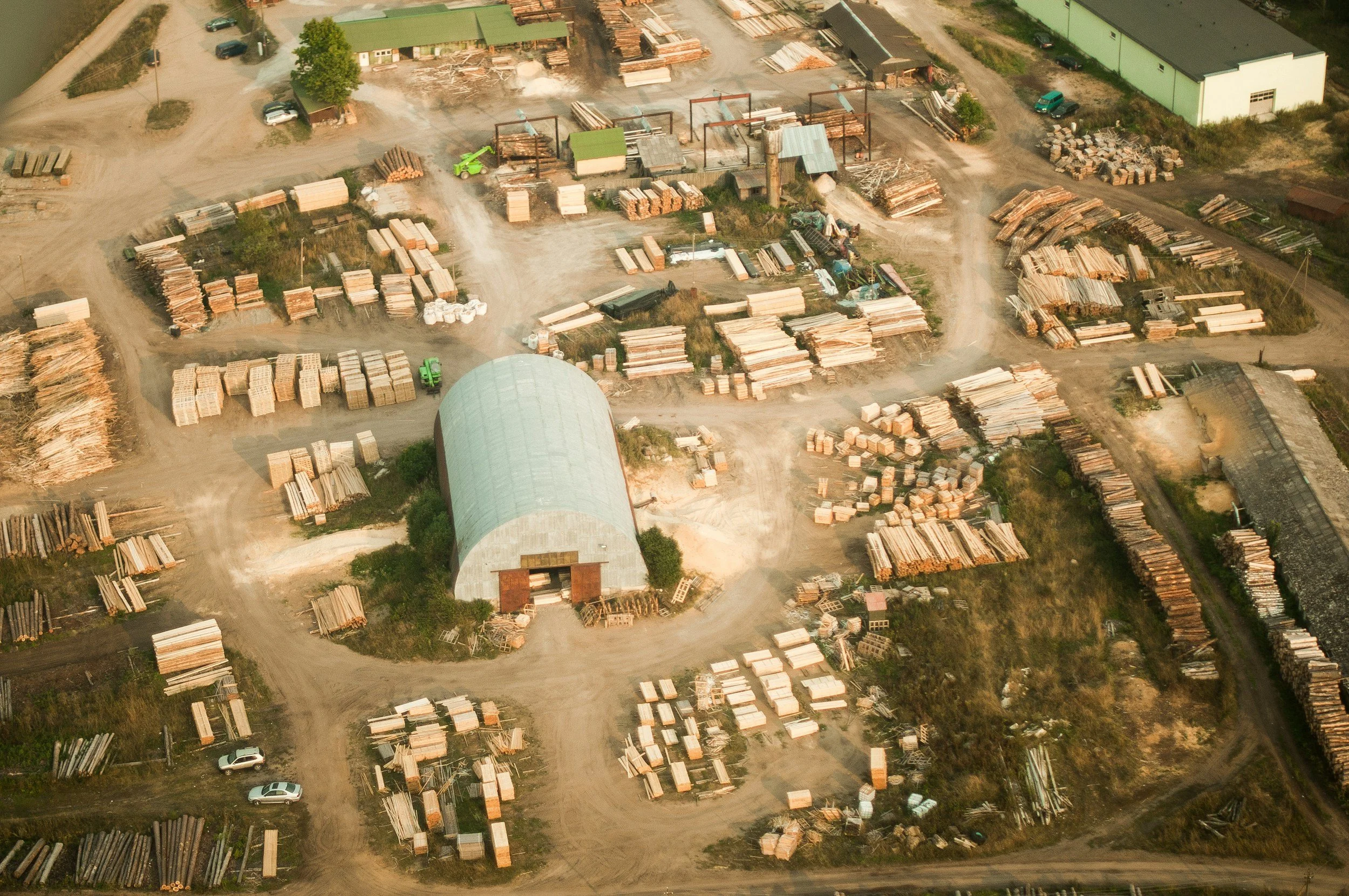 An aerial view of a lumber yard with wood stacks, vehicles, and a large warehouse building.