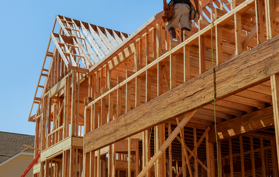 The image shows a multi-story wooden house under construction. The framing of the building is visible, with wooden beams and studs. A construction worker is on the upper level, working on the framework.