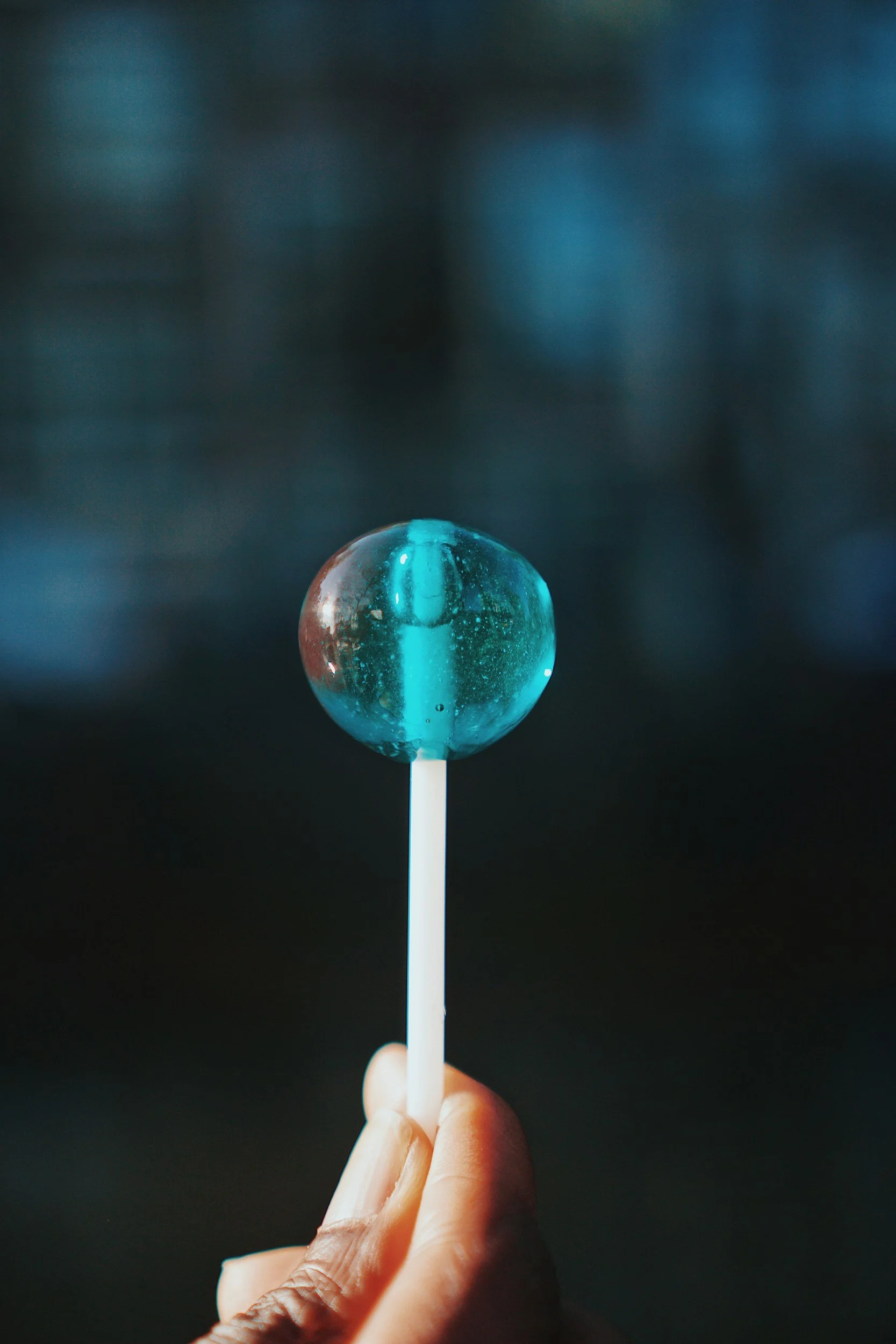 Close-up of a hand holding a blue lollipop on a stick against a dark background.