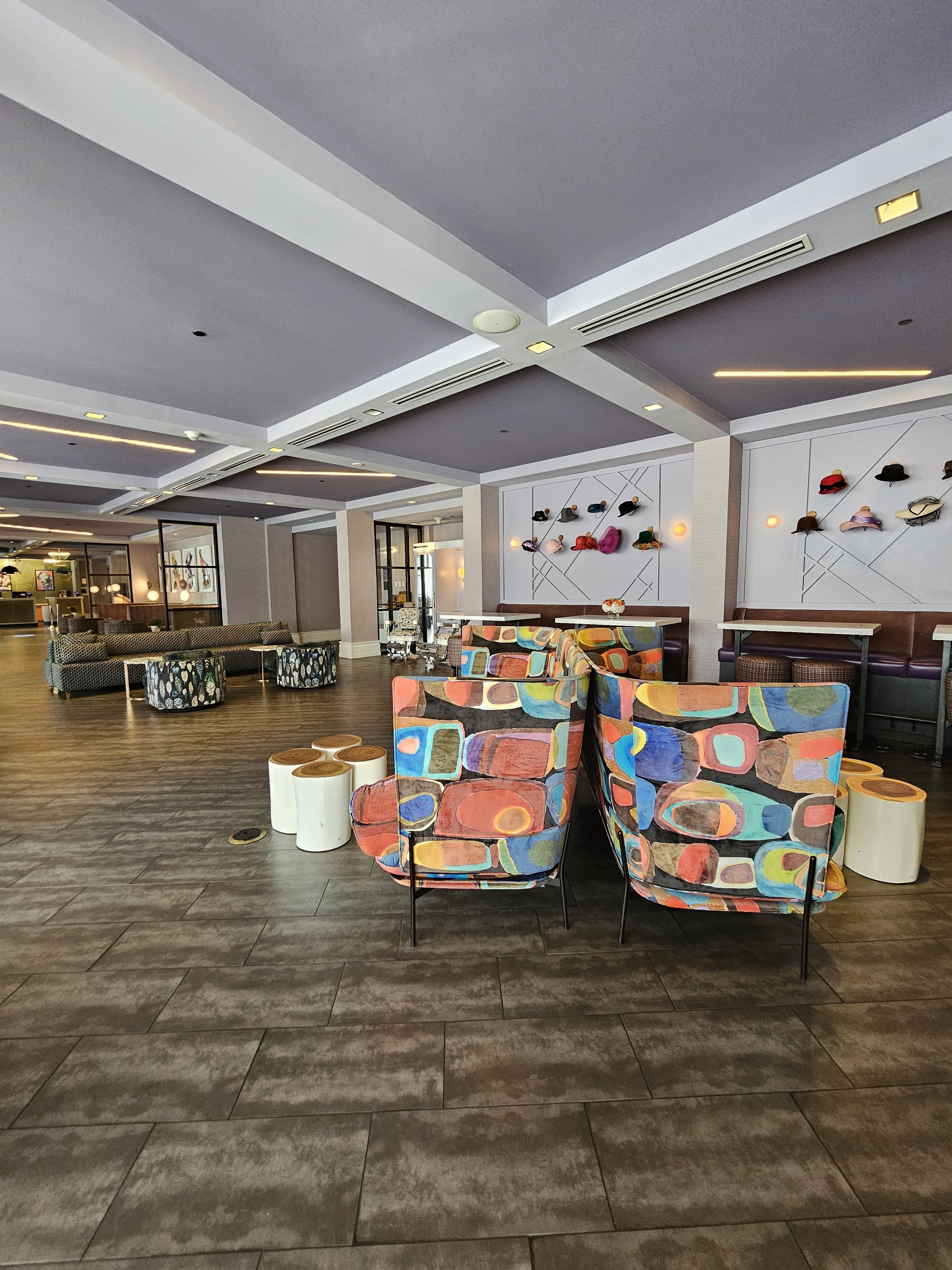 A spacious, modern lobby with colorful upholstered chairs, wall-mounted hats, and a coffee table, featuring a high ceiling with recessed lighting.