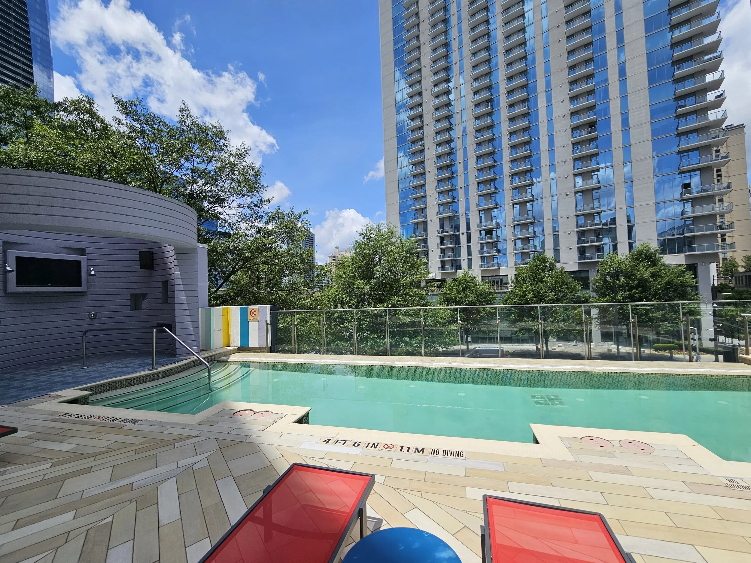 An outdoor swimming pool area in an urban setting with a high-rise building in the background, surrounded by trees and clear blue sky.