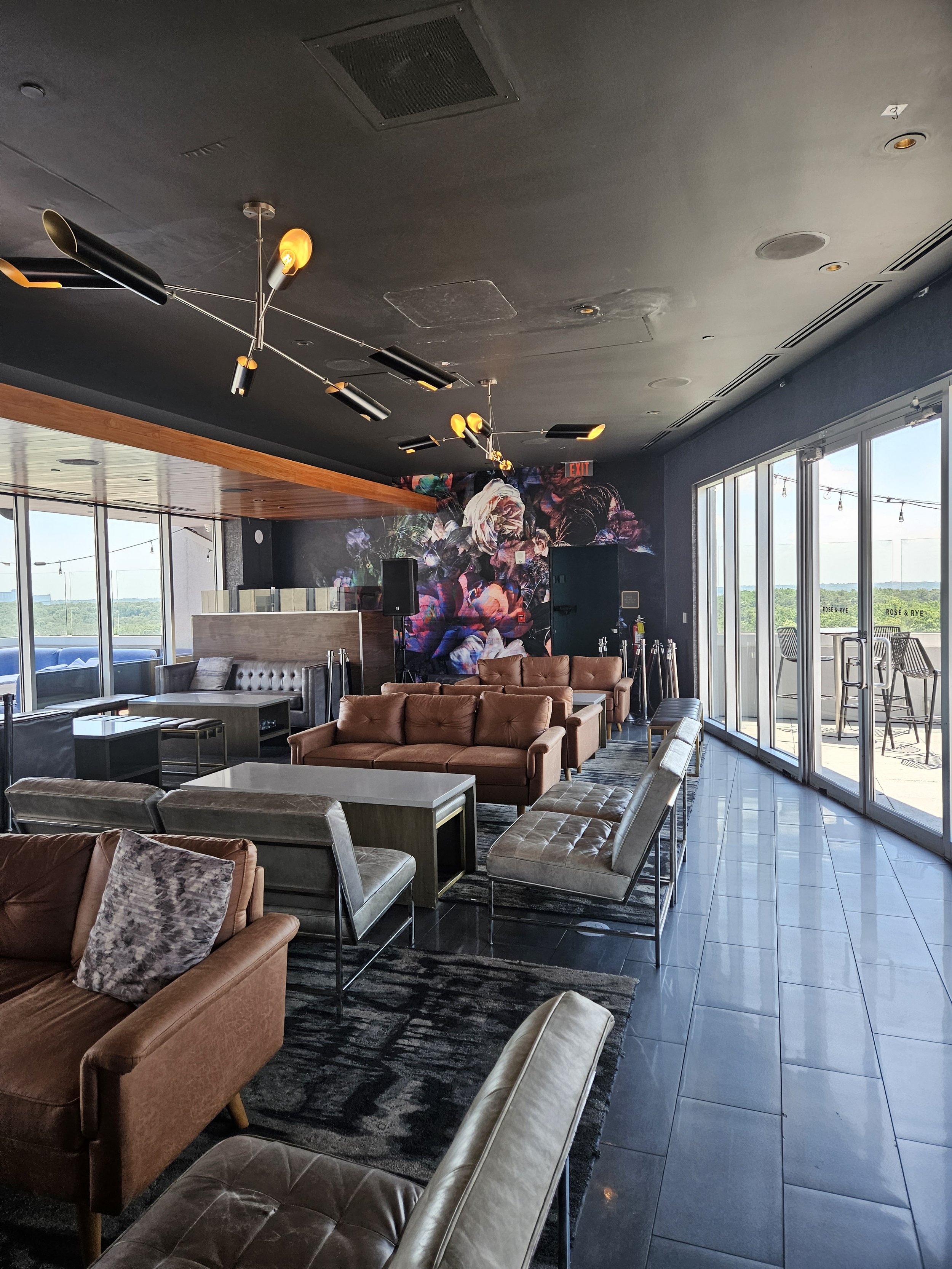 Modern lounge with leather and fabric sofas, a dark floral accent wall, large windows, and views of the outdoors. The ceiling has a contemporary black and gold lighting fixture.