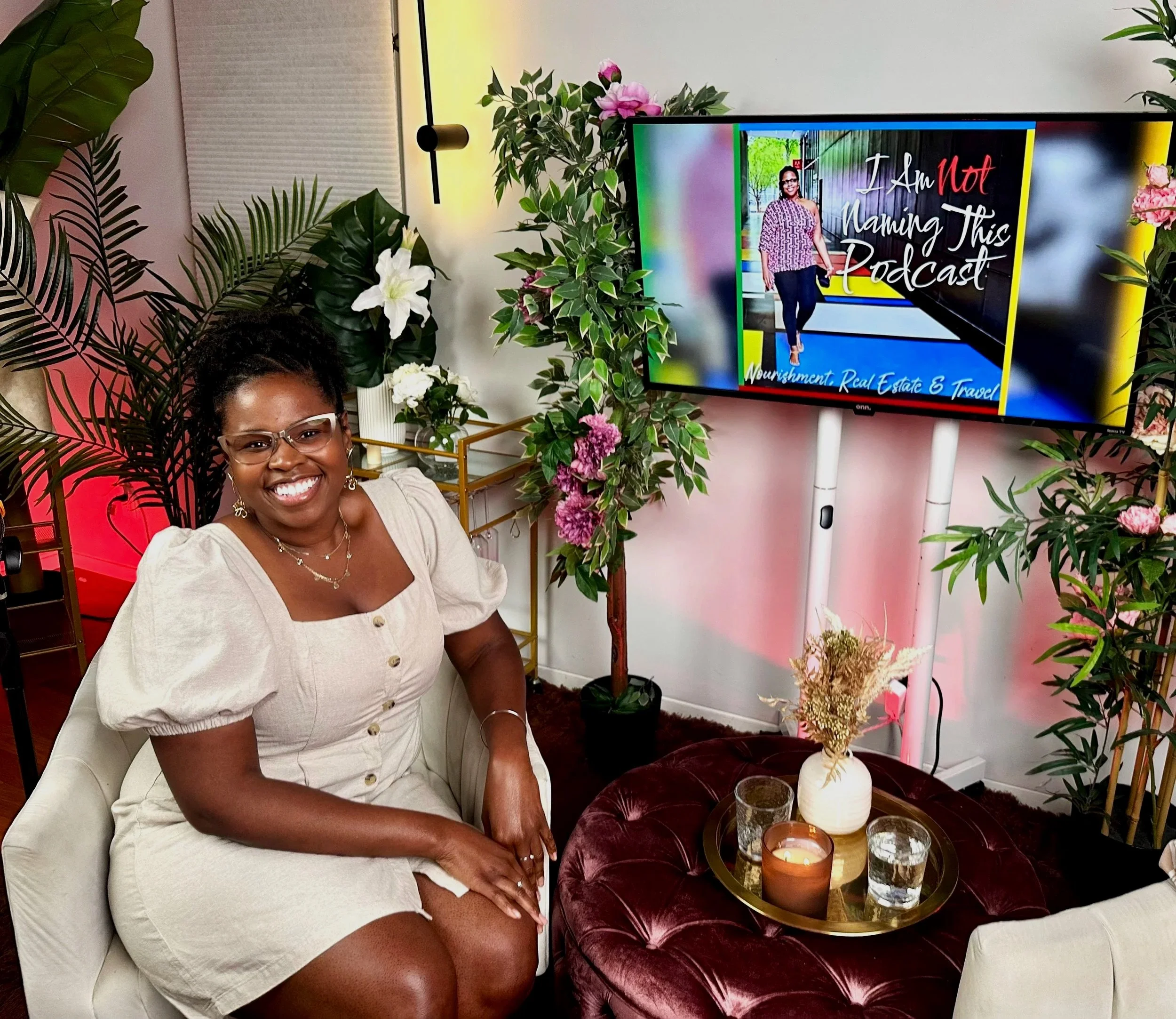 A woman sitting on a white chair surrounded by green plants and flowers, smiling at the camera, with a large screen behind her displaying a podcast titled 'I Am Not Naming This Podcast' with a picture of her standing in front of a colorful background.