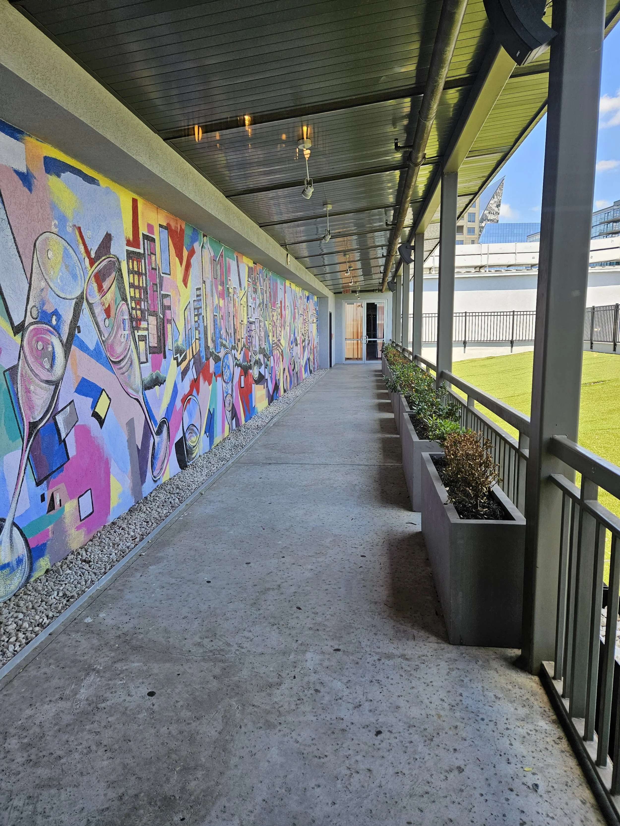 A sidewalk terrace with a mural on the wall to the left and a small lawn with plants in planters to the right. The ceiling is painted black with lighting fixtures, and the terrace has a railing on the right side, with buildings visible in the background.