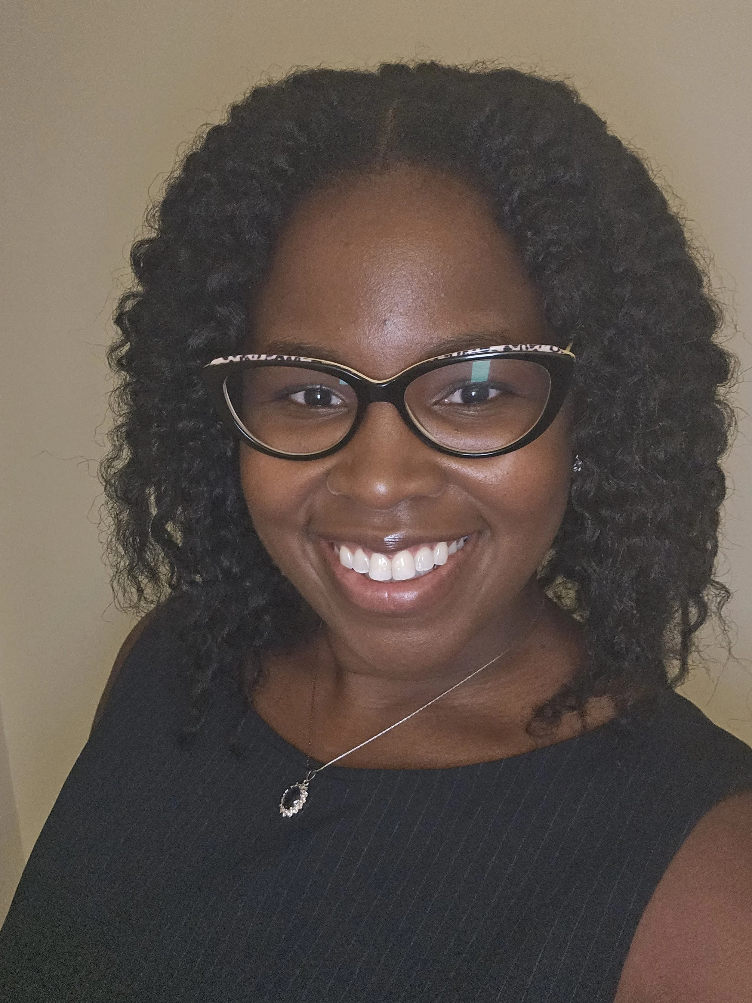 Chernice Dieudonne - A woman with curly black hair, wearing glasses, smiling, dressed in a black top, with a necklace, and a beige background.