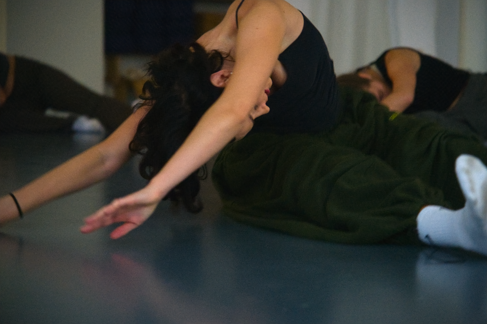 A woman practicing yoga stretches on a mat in a dimly lit room, wearing a black tank top, camo pants, and white sneakers.