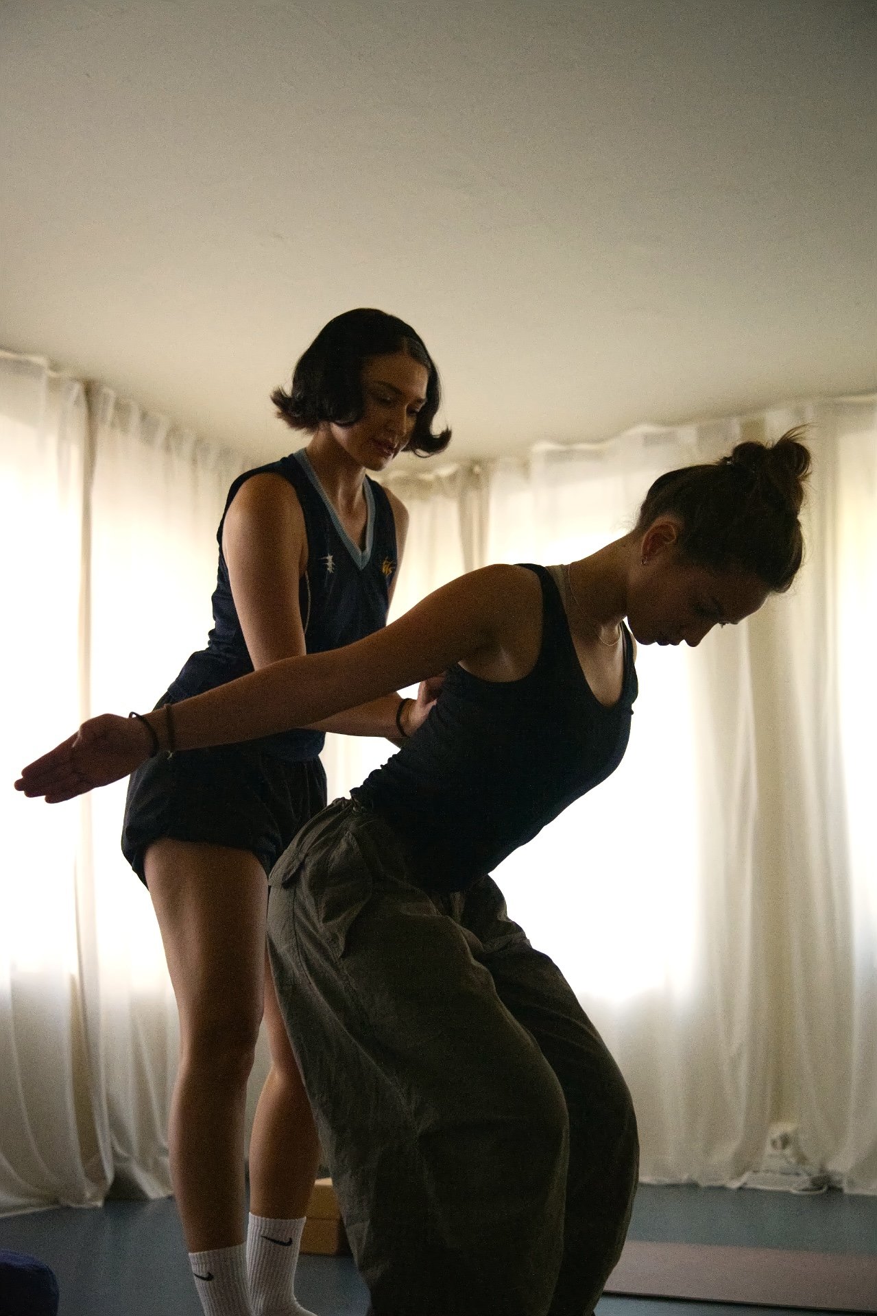 Two young women practicing physical therapy exercises in a room with natural light curtains. One woman is assisting the other, who is bending forward with her arms extended back.