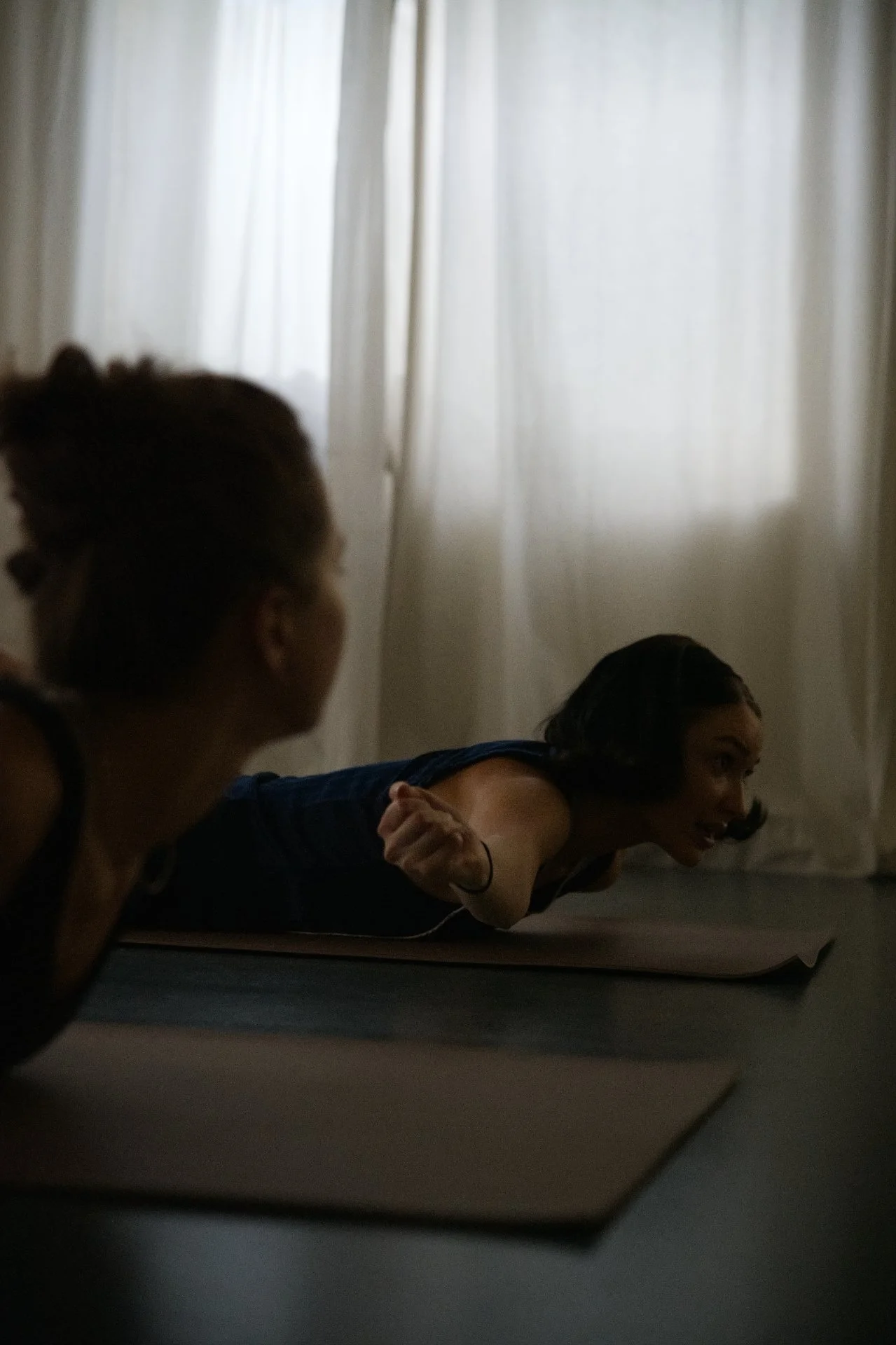 Two women practicing yoga or stretching indoors, with light coming through curtains in the background.