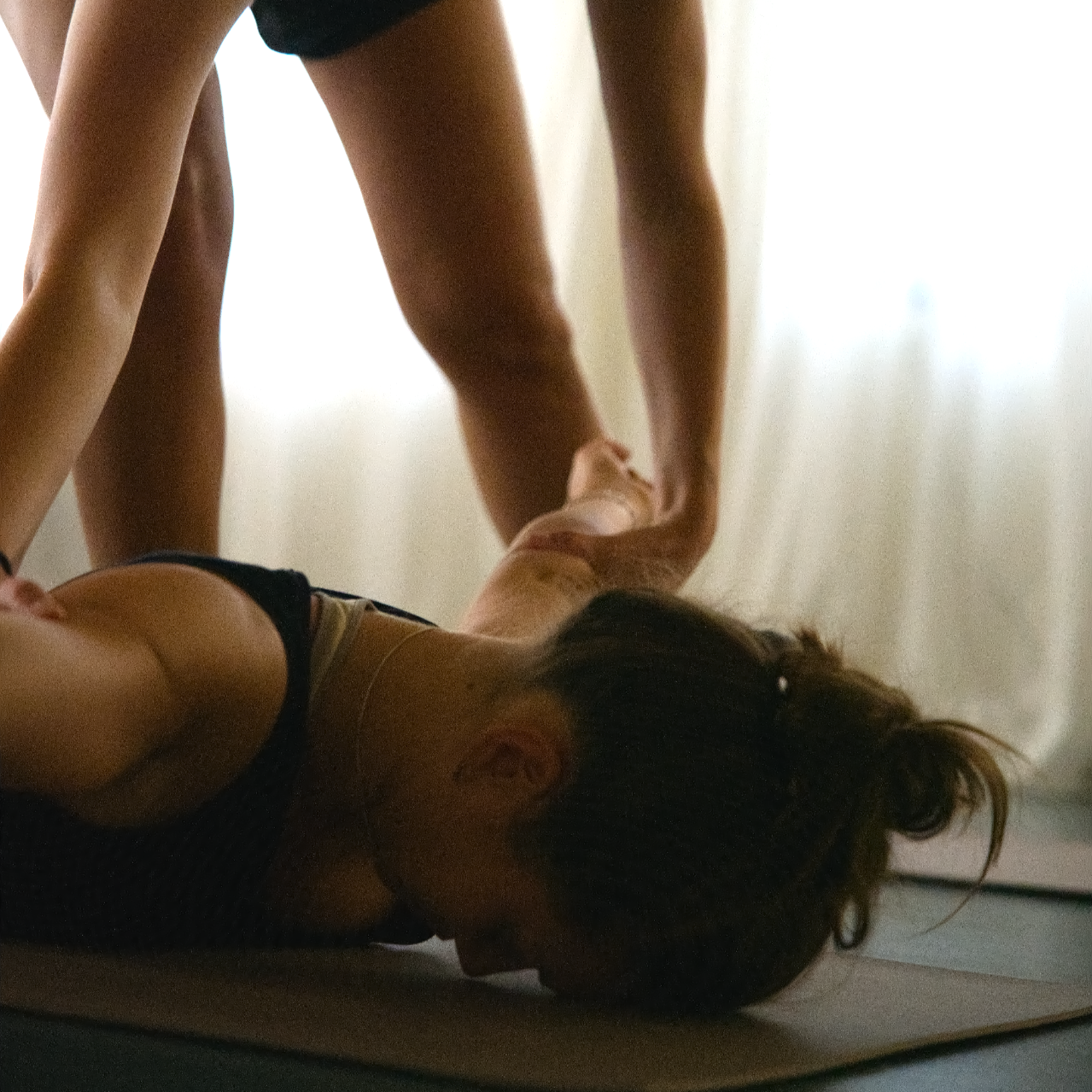A woman lying on a yoga mat with her head turned to the side, a person standing over her holding her hand, in a brightly lit room with sheer curtains.
