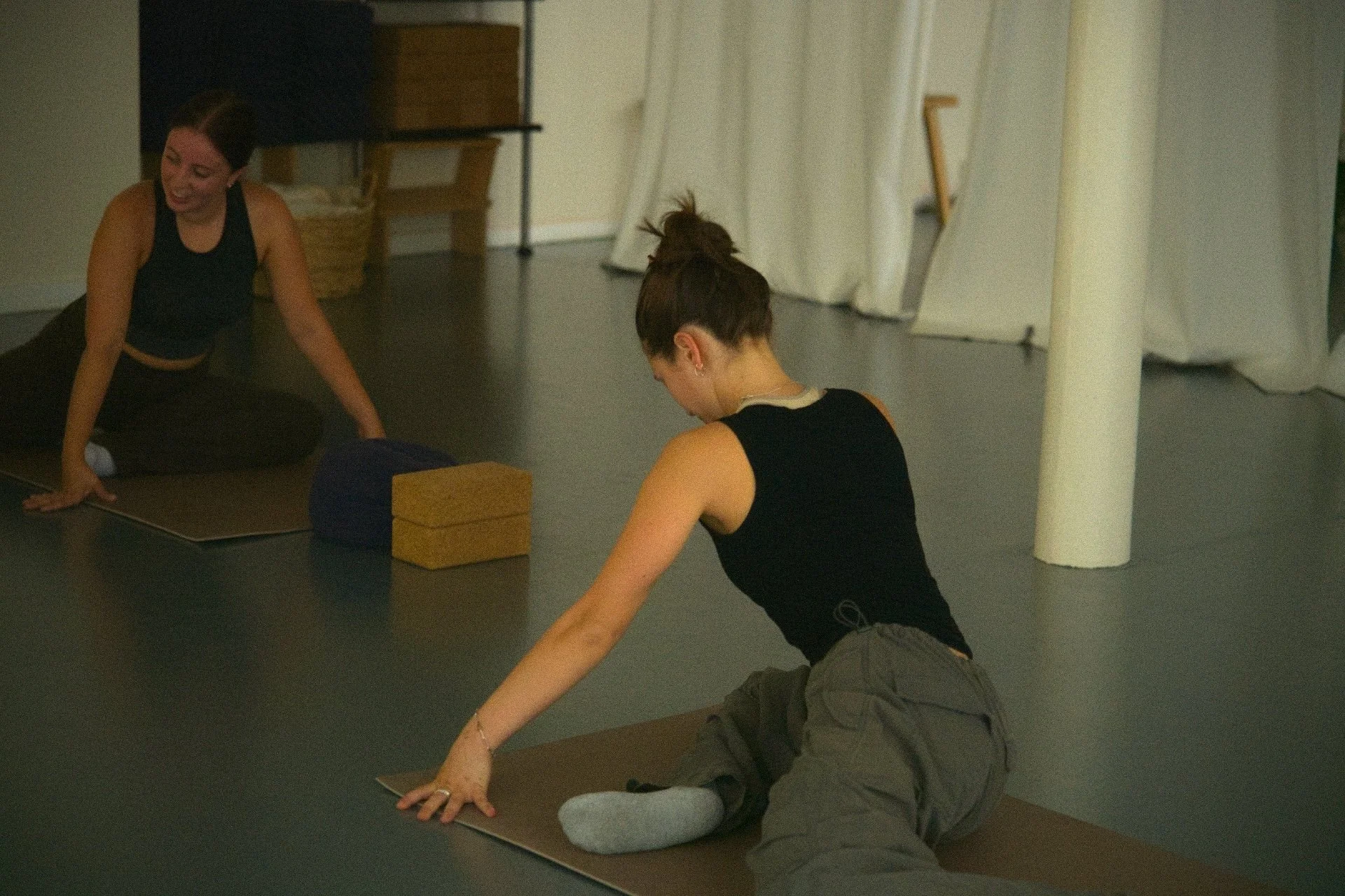 Two women practicing yoga or stretching on yoga mats in a studio. One woman is sitting on her knees with palms flat on the floor, wearing gray pants and a black sleeveless top. The other woman is sitting on her heels, smiling, wearing black pants and a black sleeveless top.