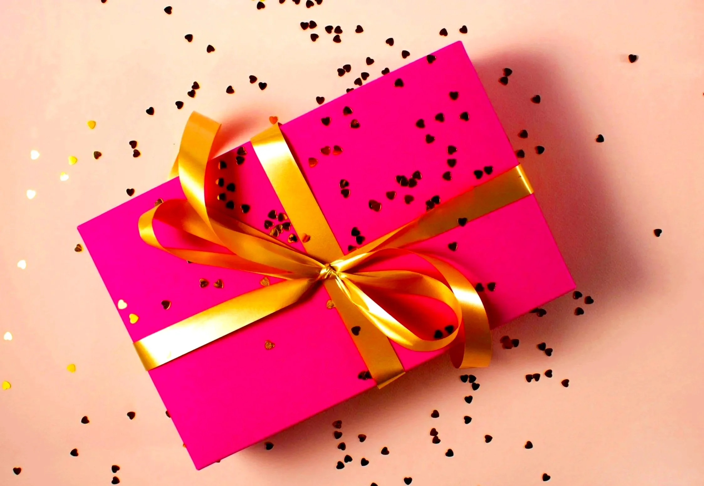 Pink gift box with a gold ribbon and bow, surrounded by black and gold heart-shaped confetti on a pink background.