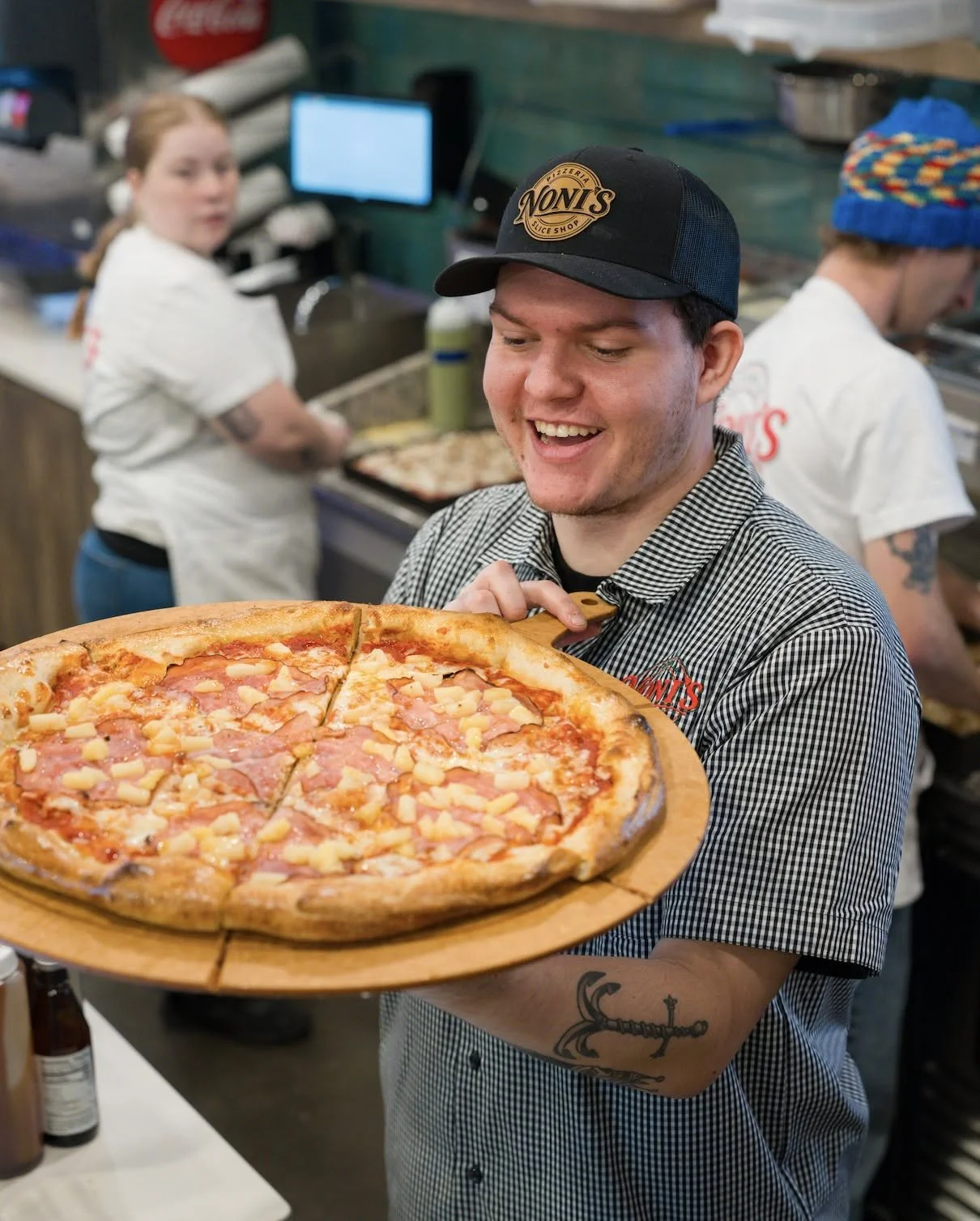 A man smiling and holding a pizza on a wooden tray inside a pizzeria, with two employees in the background preparing food.