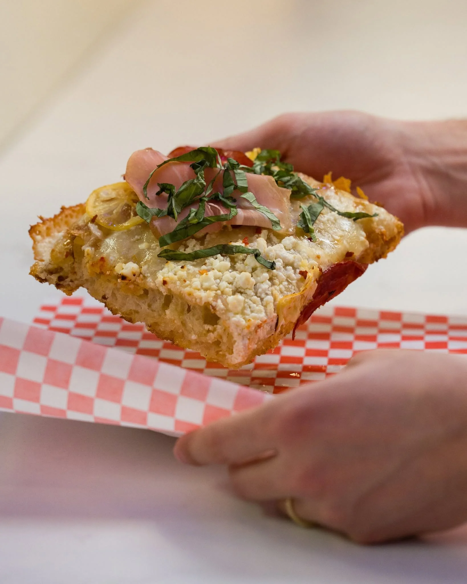 A close-up of a slice of pizza topped with herbs, ham, and yellow bell peppers, held by a person's hand against a white background.